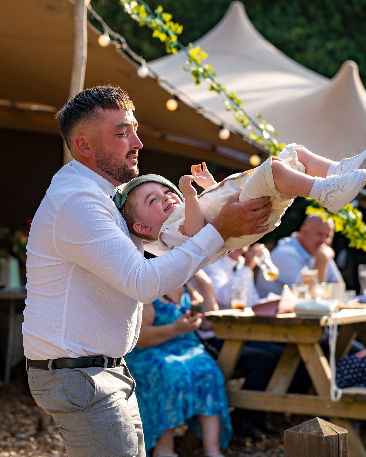 A man in a white shirt playfully lifts a laughing baby at an outdoor wedding reception under a tent.