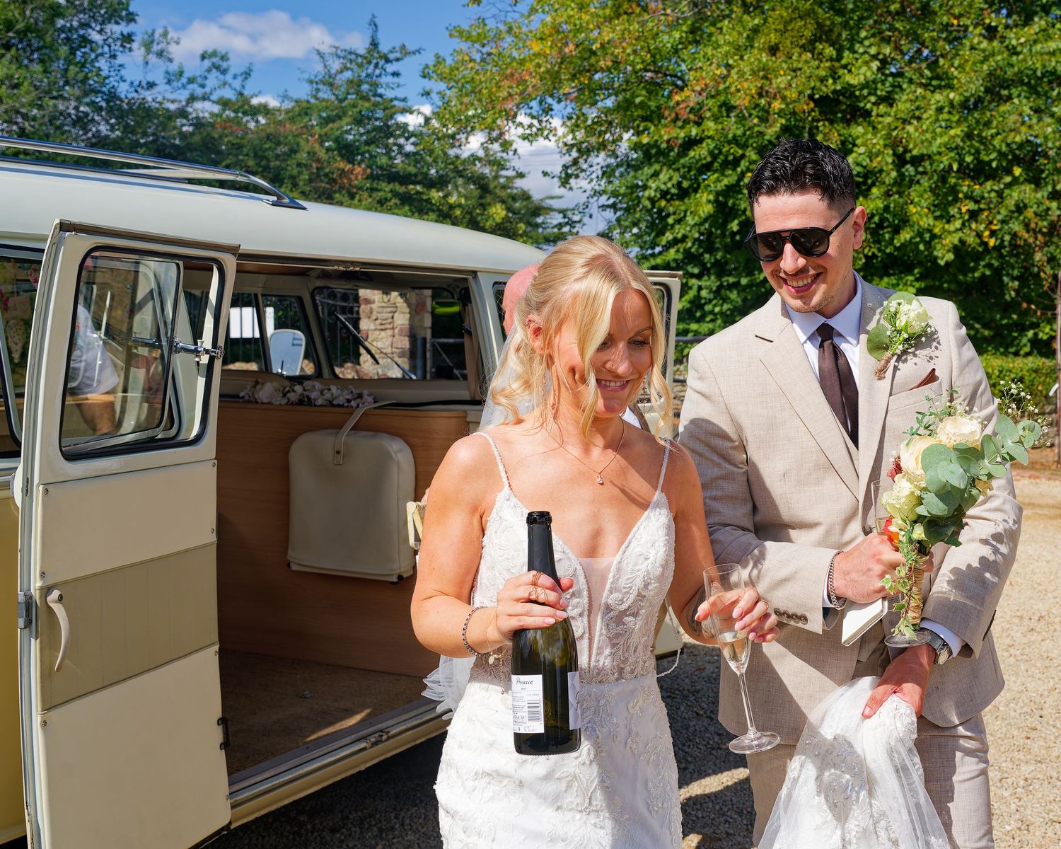 A bride and groom celebrate beside a vintage VW campervan, bride holding a wine bottle outdoors.