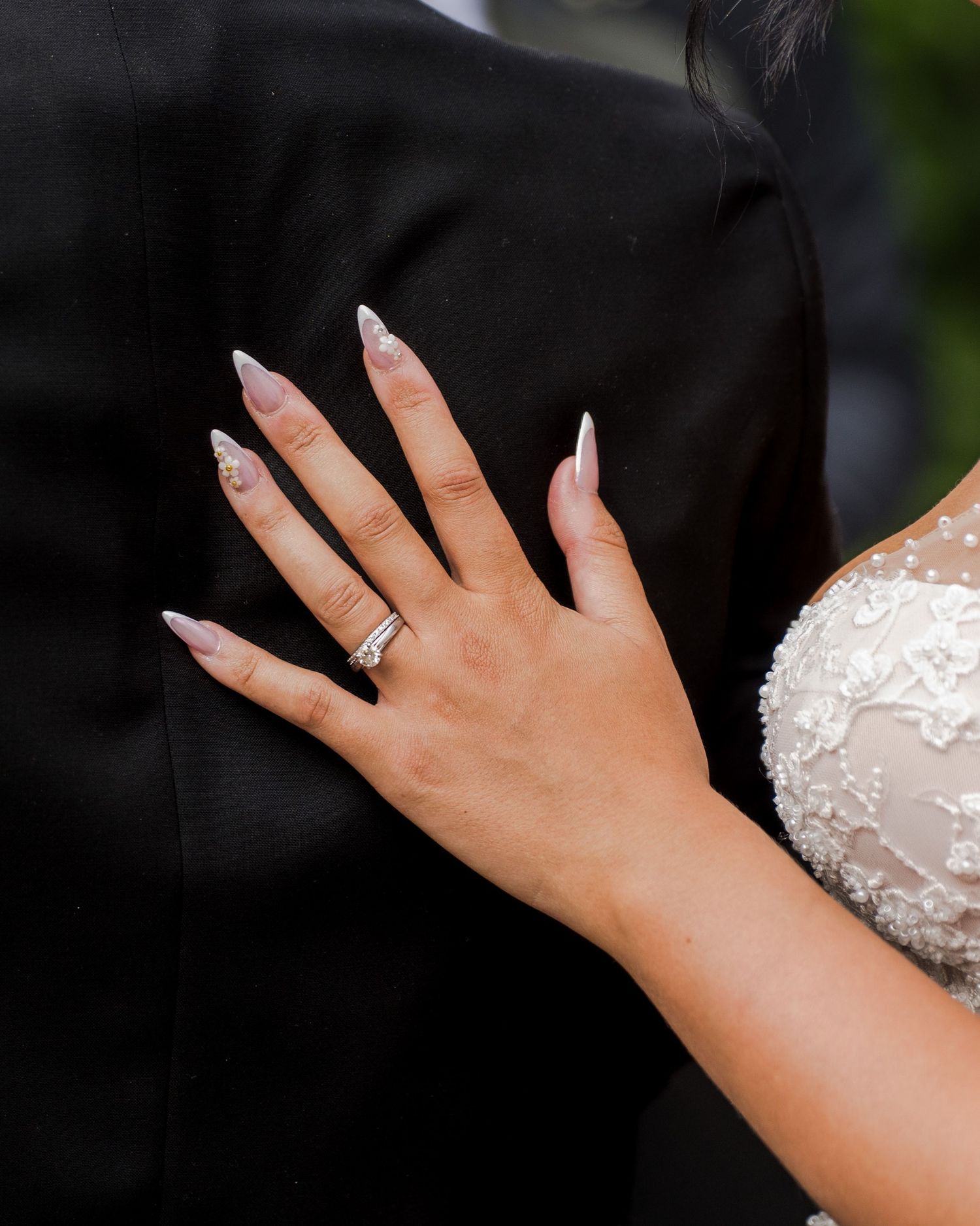 Bride displays diamond wedding ring with white almond-shaped nails against grooms dark suit jacket.