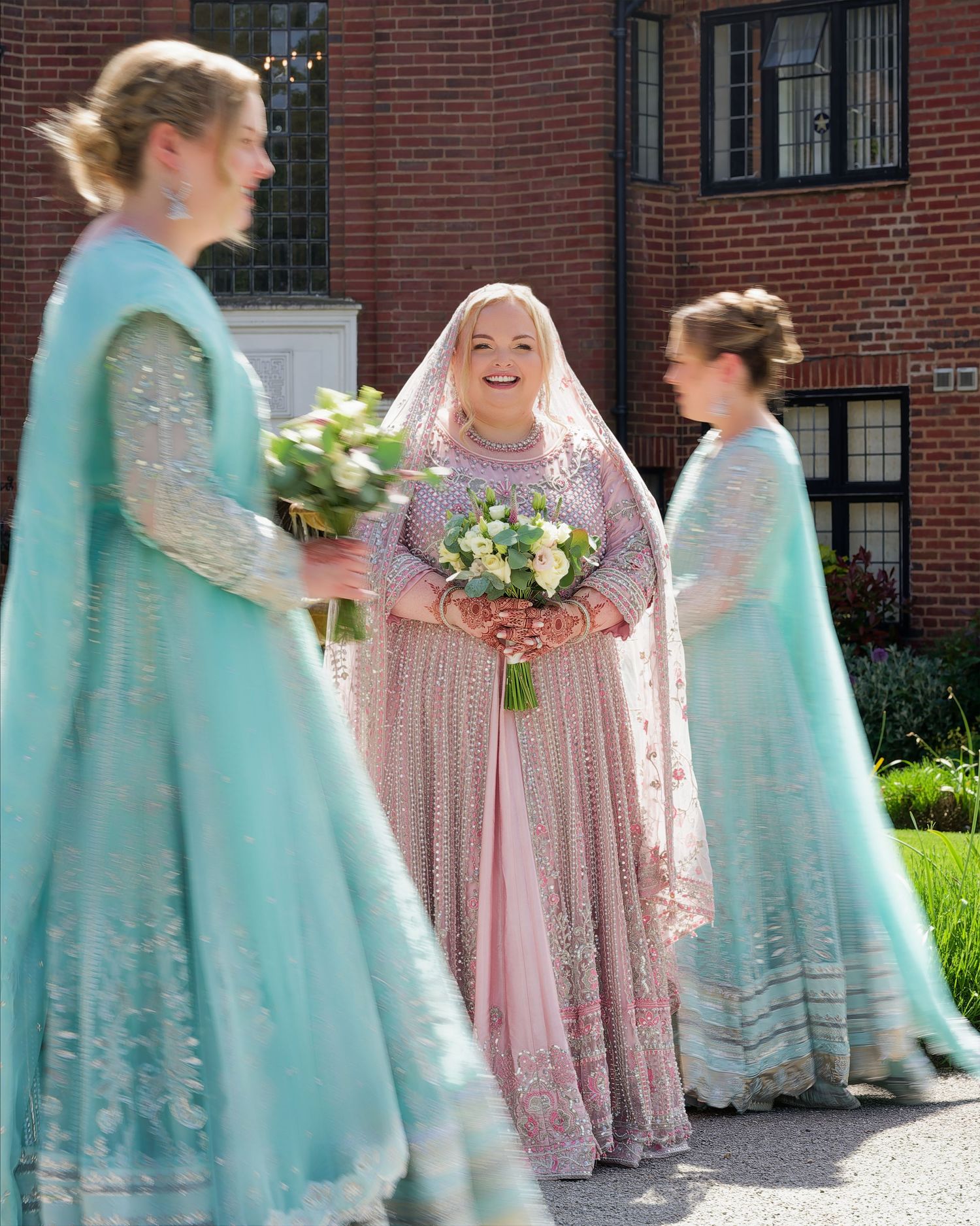 Radiant bride in pink embroidered gown smiles between bridesmaids in teal outfits outdoors.
