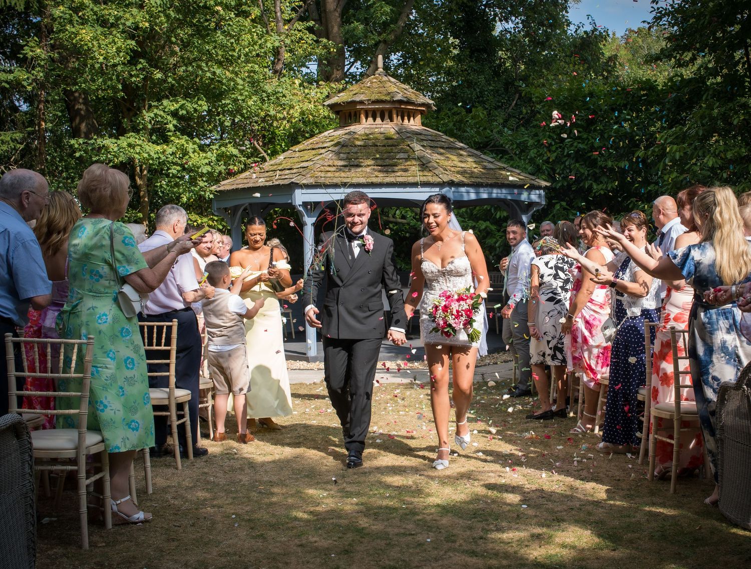 Bride and groom walk down outdoor aisle as guests throw confetti at a garden wedding ceremony.