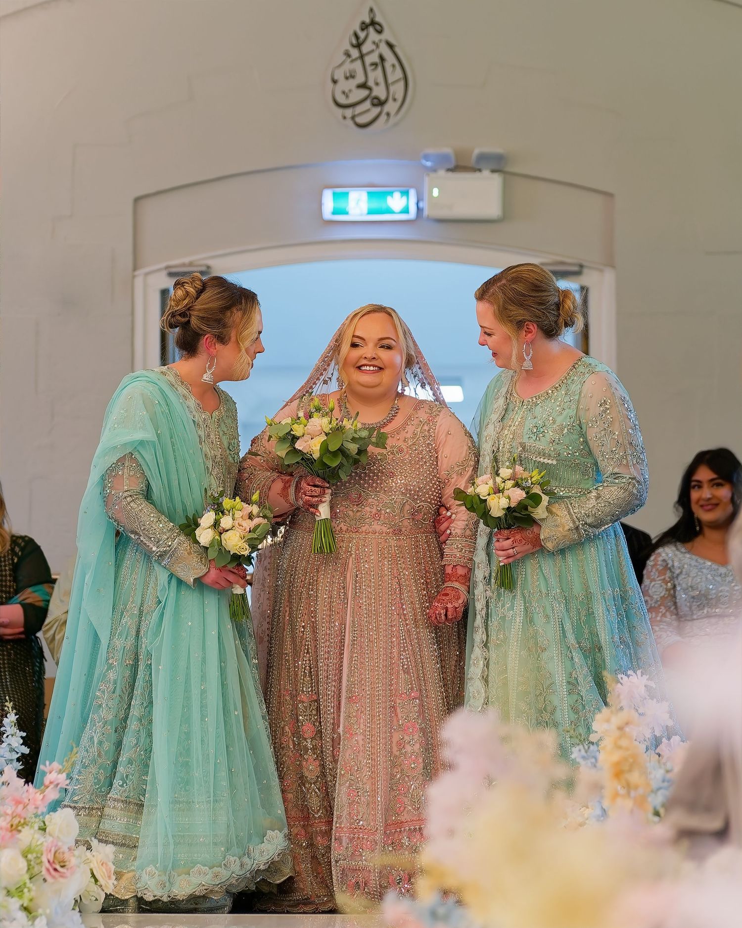 Bride in pink embroidered gown flanked by bridesmaids in turquoise at wedding ceremony.