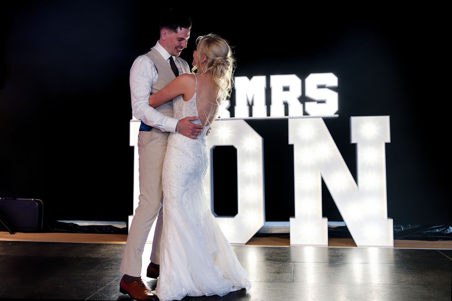 Bride and groom dancing together in front of illuminated MRS ON letter signs at their wedding.
