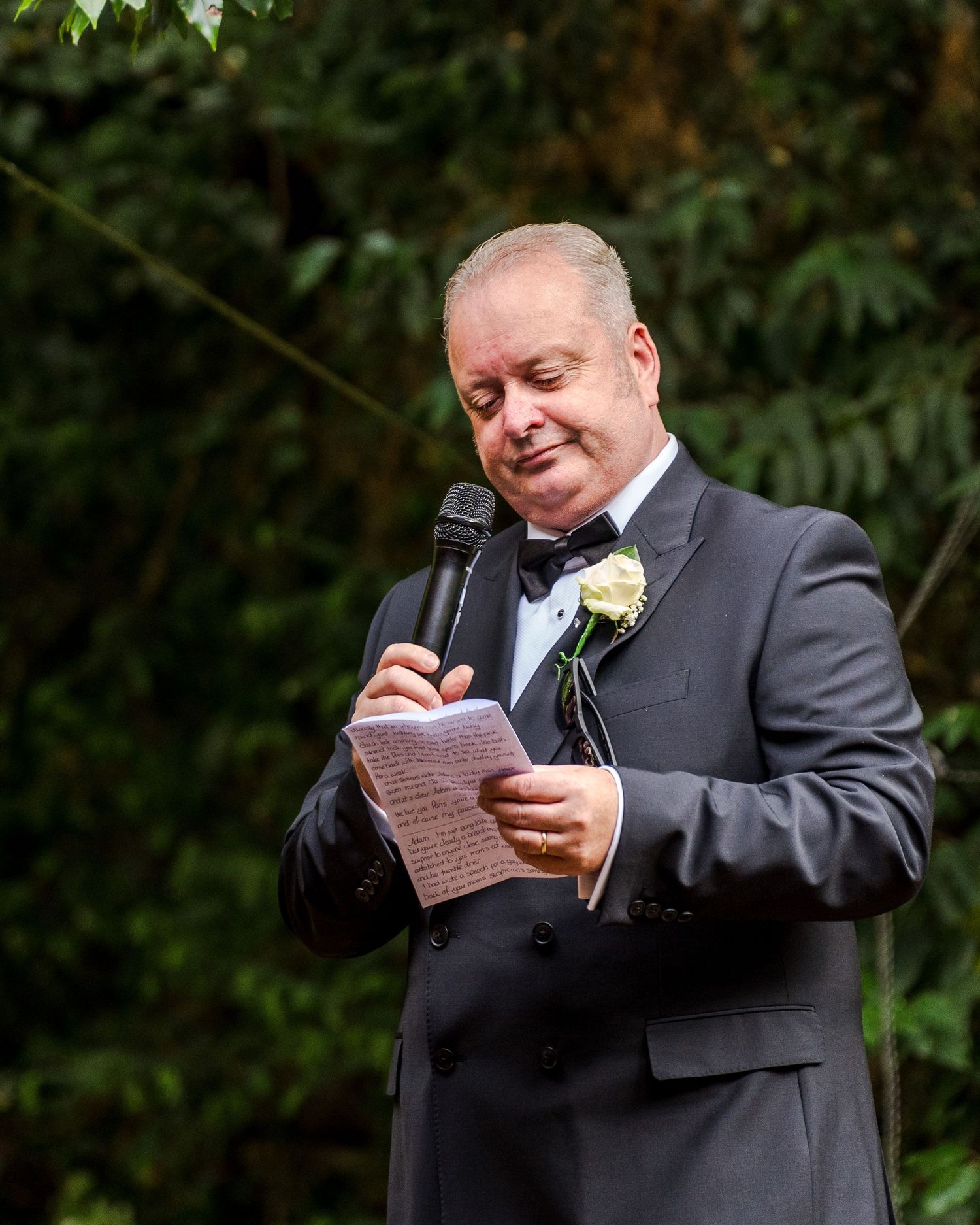 Officiant in dark suit reads handwritten notes at outdoor wedding ceremony surrounded by greenery.