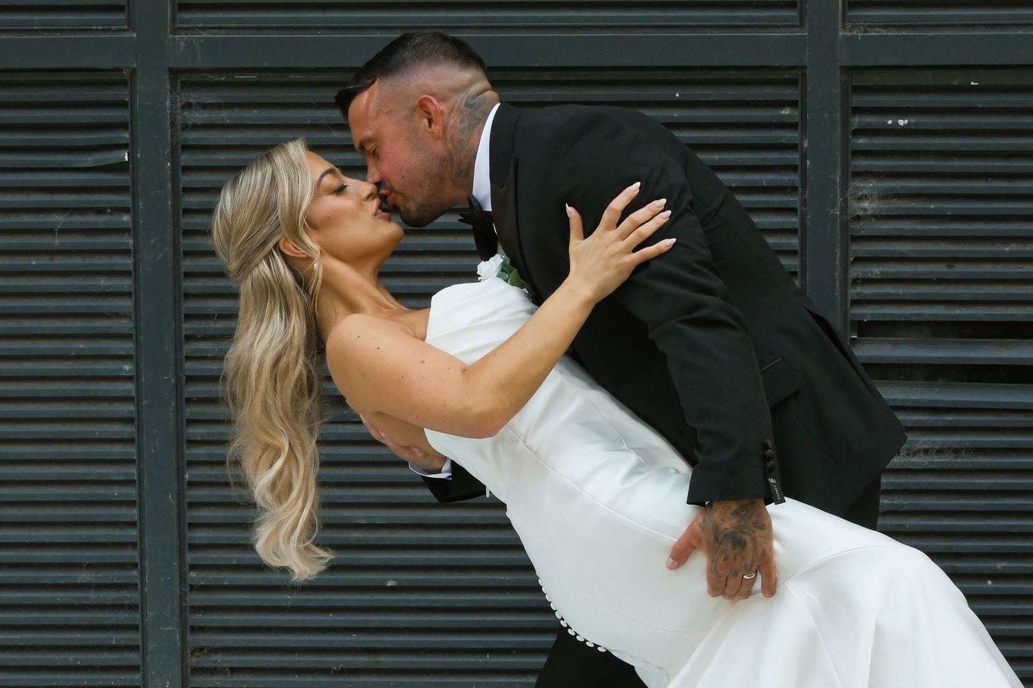 Bride and groom share a romantic dip kiss in front of a dark striped wall on their wedding day.