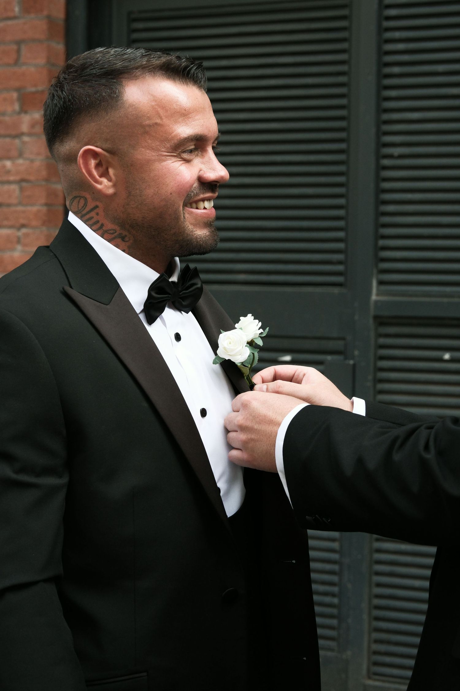 Smiling groom in black tuxedo with bow tie has white rose boutonniere pinned to lapel.