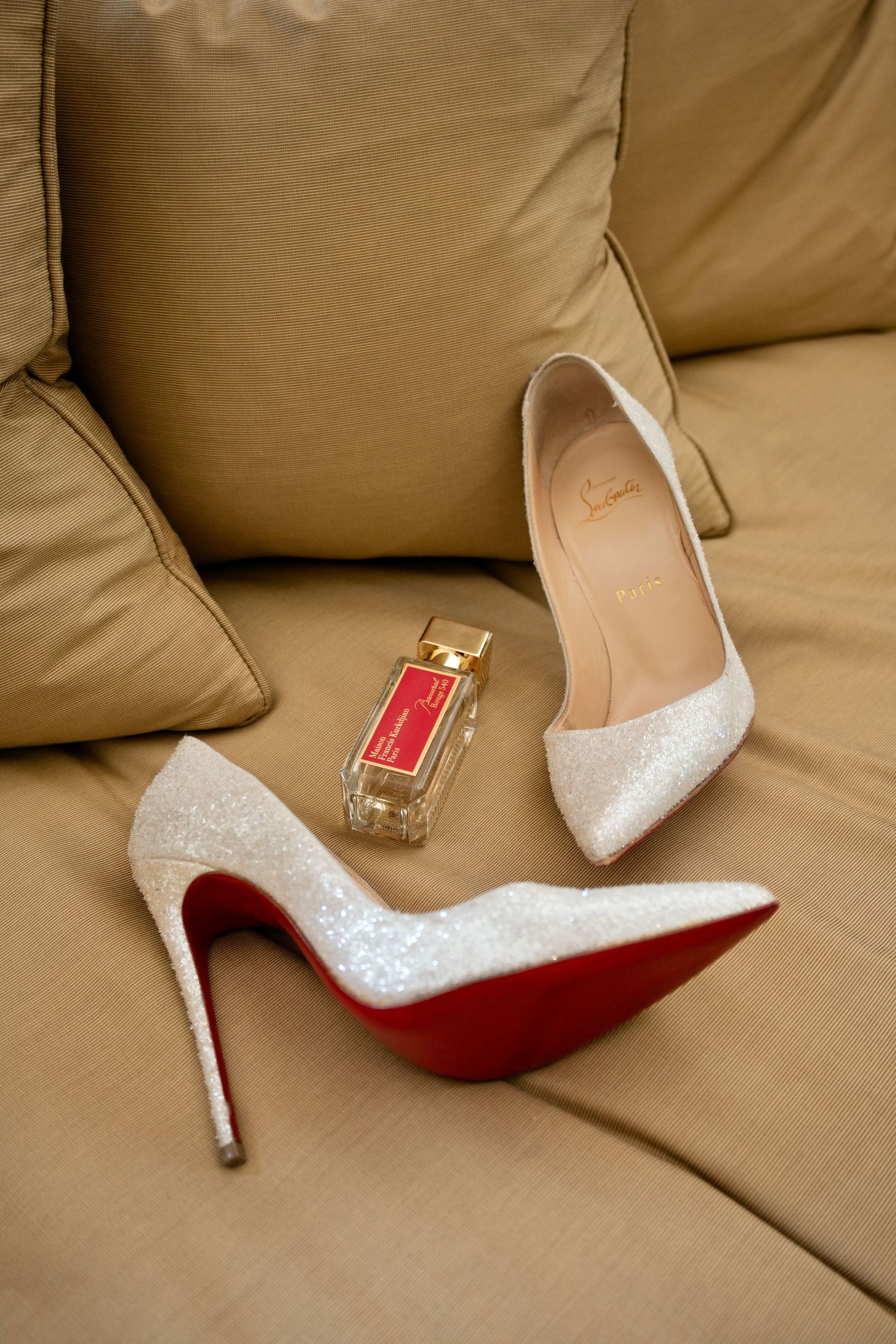 Sparkling white Christian Louboutin stiletto heels with red soles beside luxury perfume bottle on beige sofa.