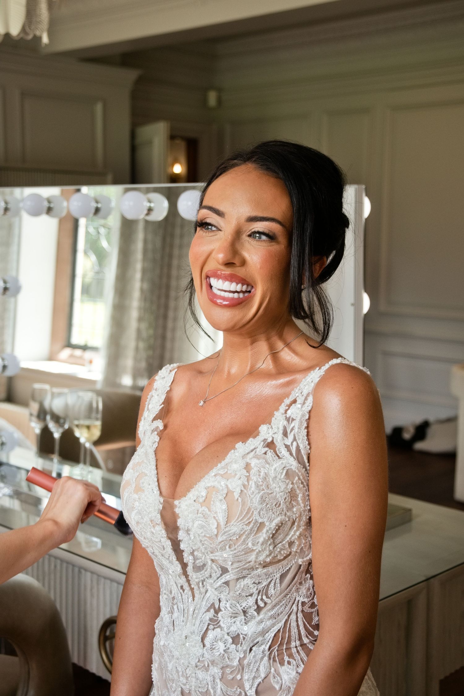 Radiant bride with dark updo smiles brightly in deep V-neck lace wedding gown at Hollywood vanity mirror.