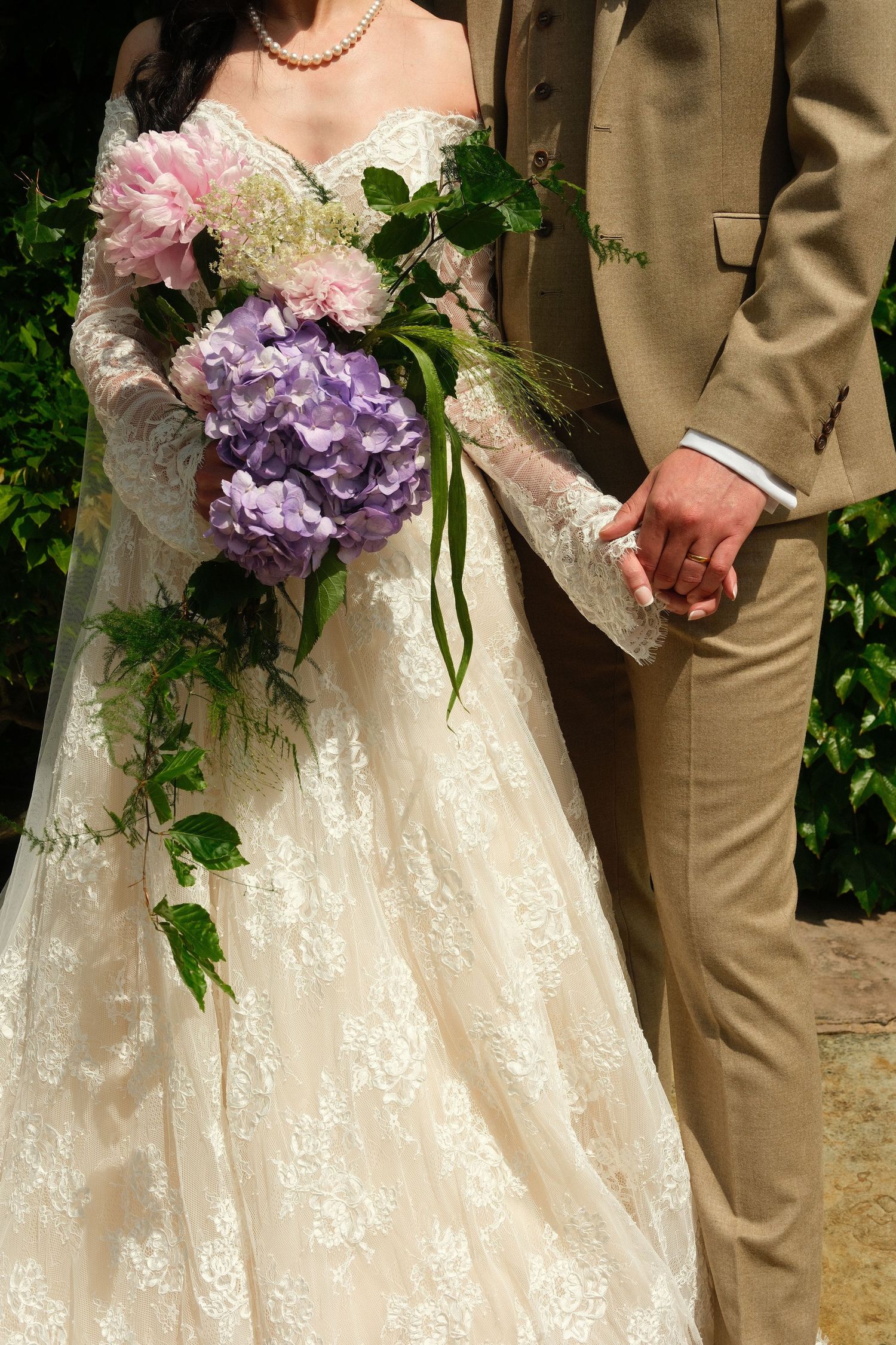 Bride in lace gown holds purple hydrangea bouquet while groom in tan suit holds her hand outdoors.