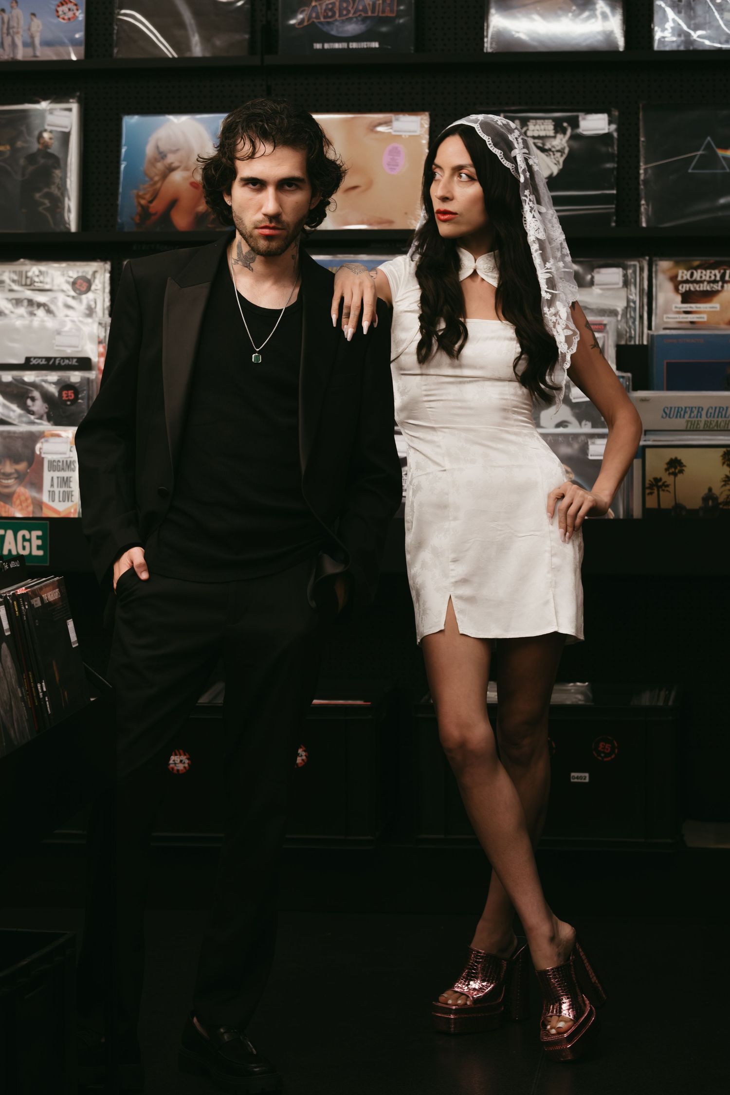 A man in black and woman in white mini dress and veil pose together in a vinyl record store.