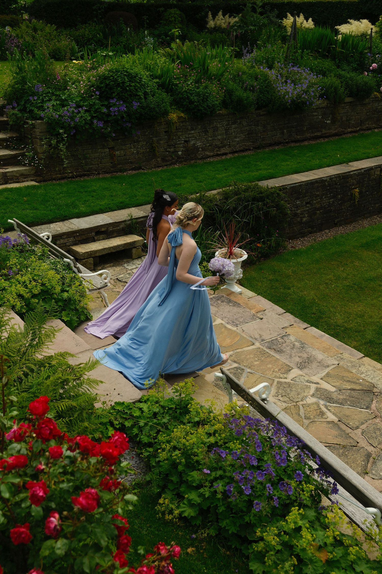 Two bridesmaids in blue and lavender gowns walking through a terraced English garden.