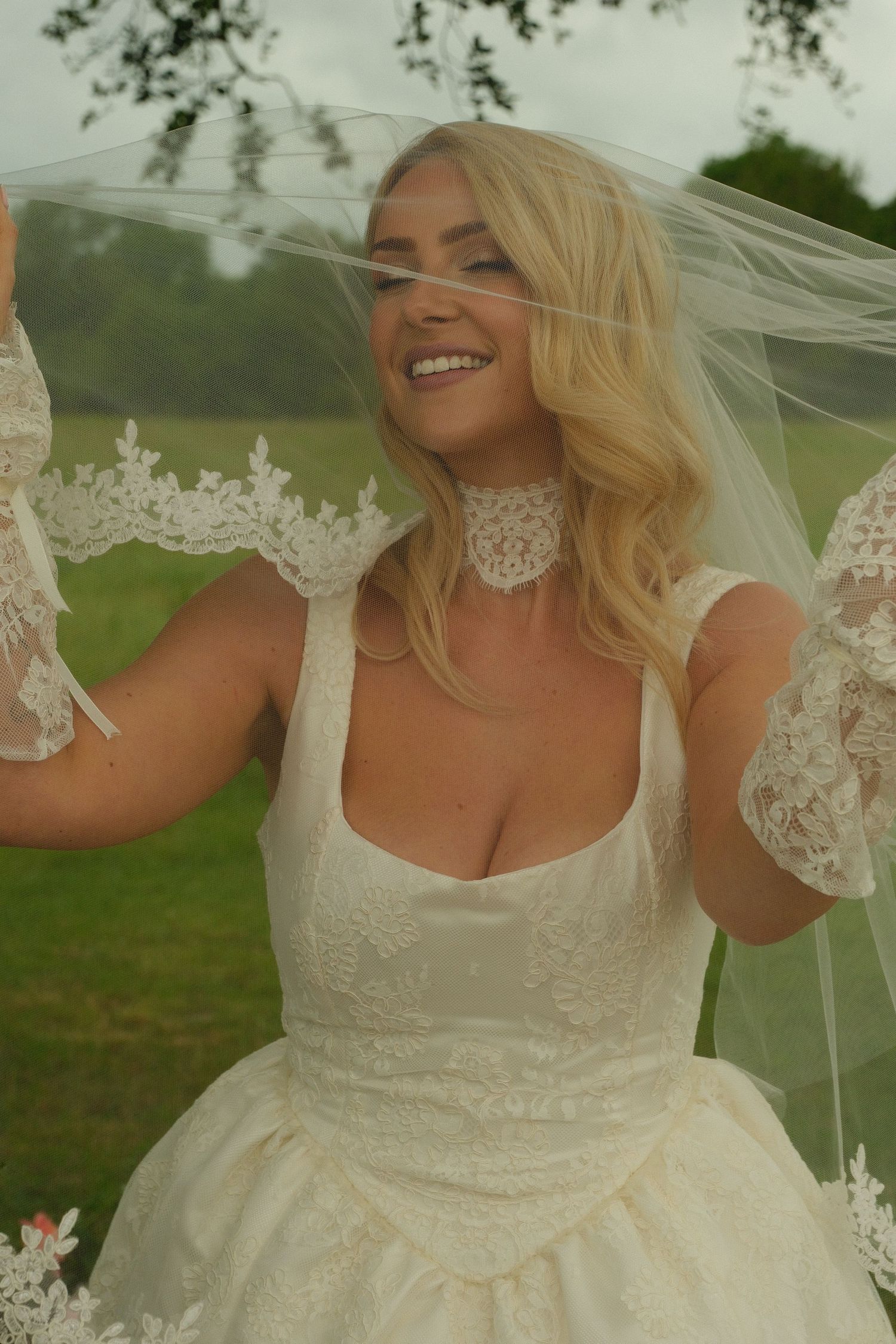 Joyful blonde bride in vintage lace wedding dress lifting veil outdoors in green field.