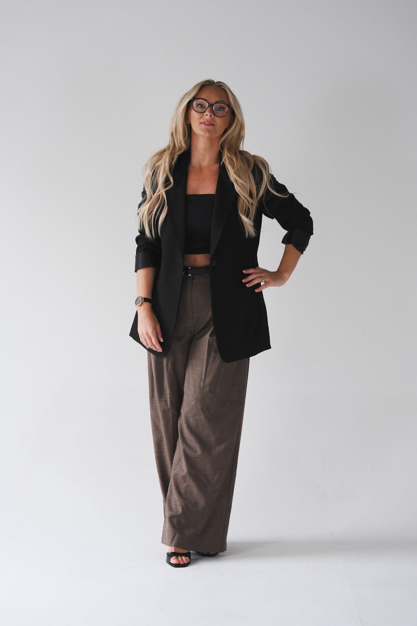 Blonde woman in black blazer, crop top, and wide-leg brown trousers posing confidently in a studio setting.