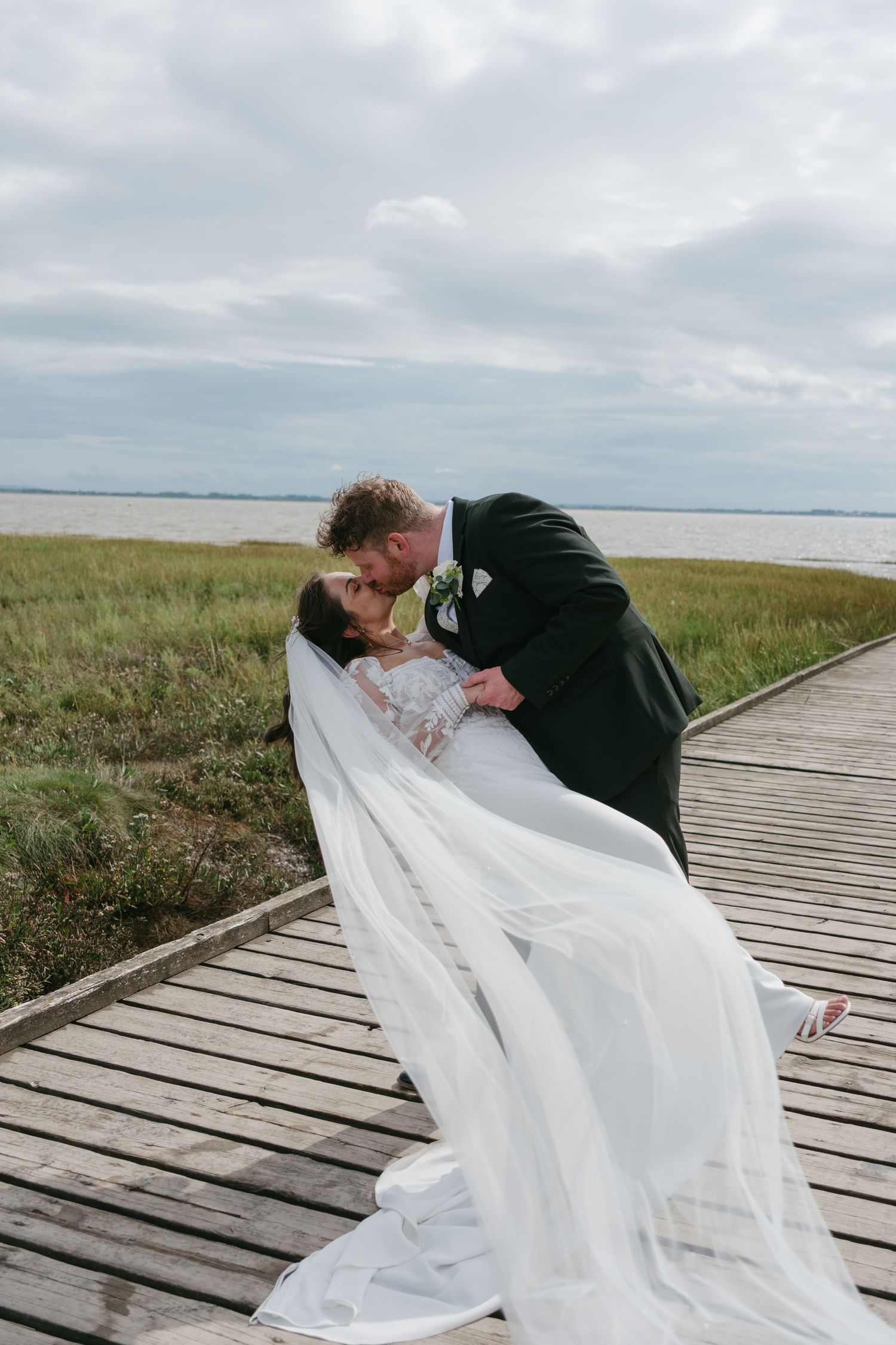 Bride and groom sharing a romantic dip kiss on a wooden boardwalk near a coastal marsh on their wedding day.