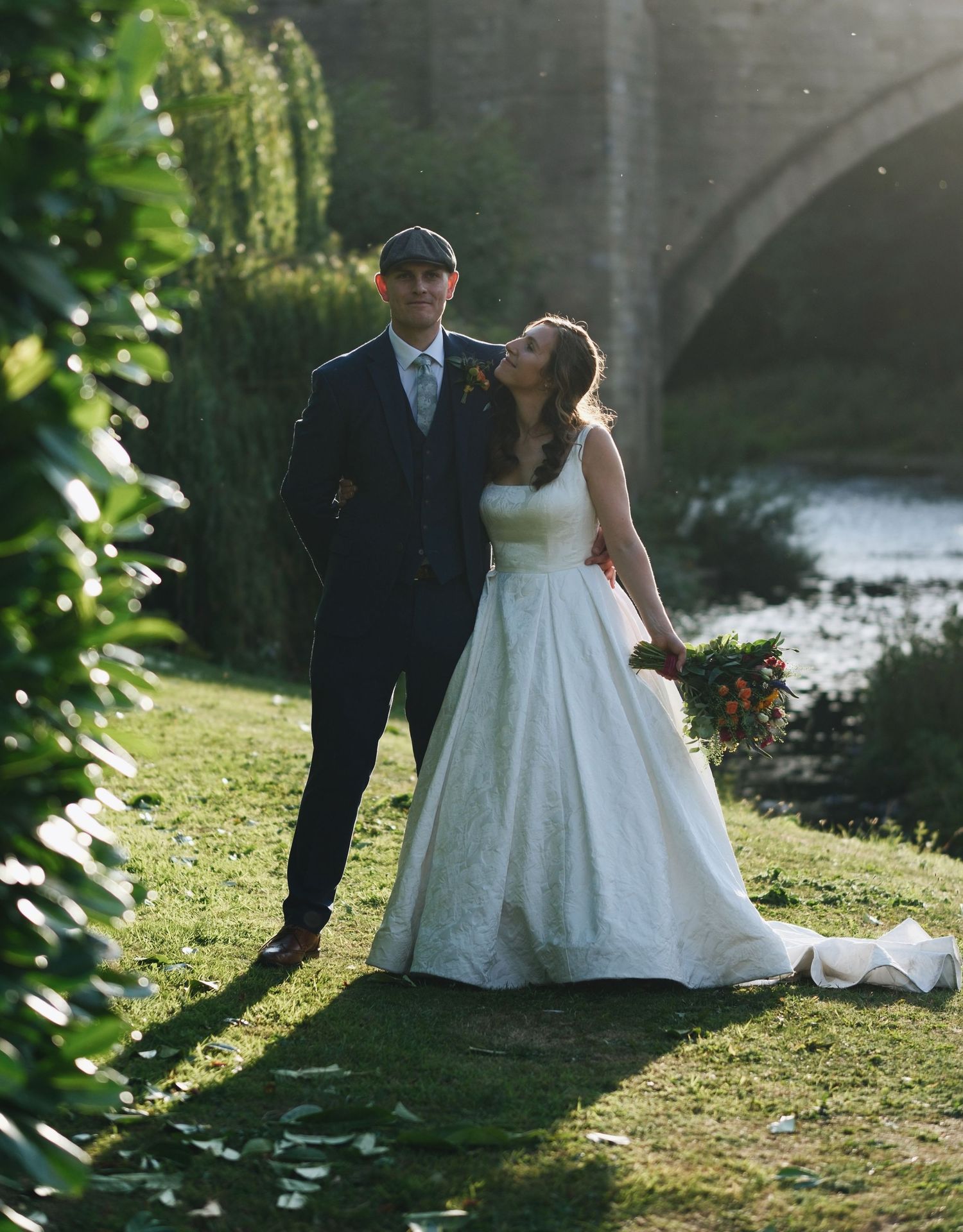 Bride and groom walking by a river with a stone bridge backdrop on their wedding day.