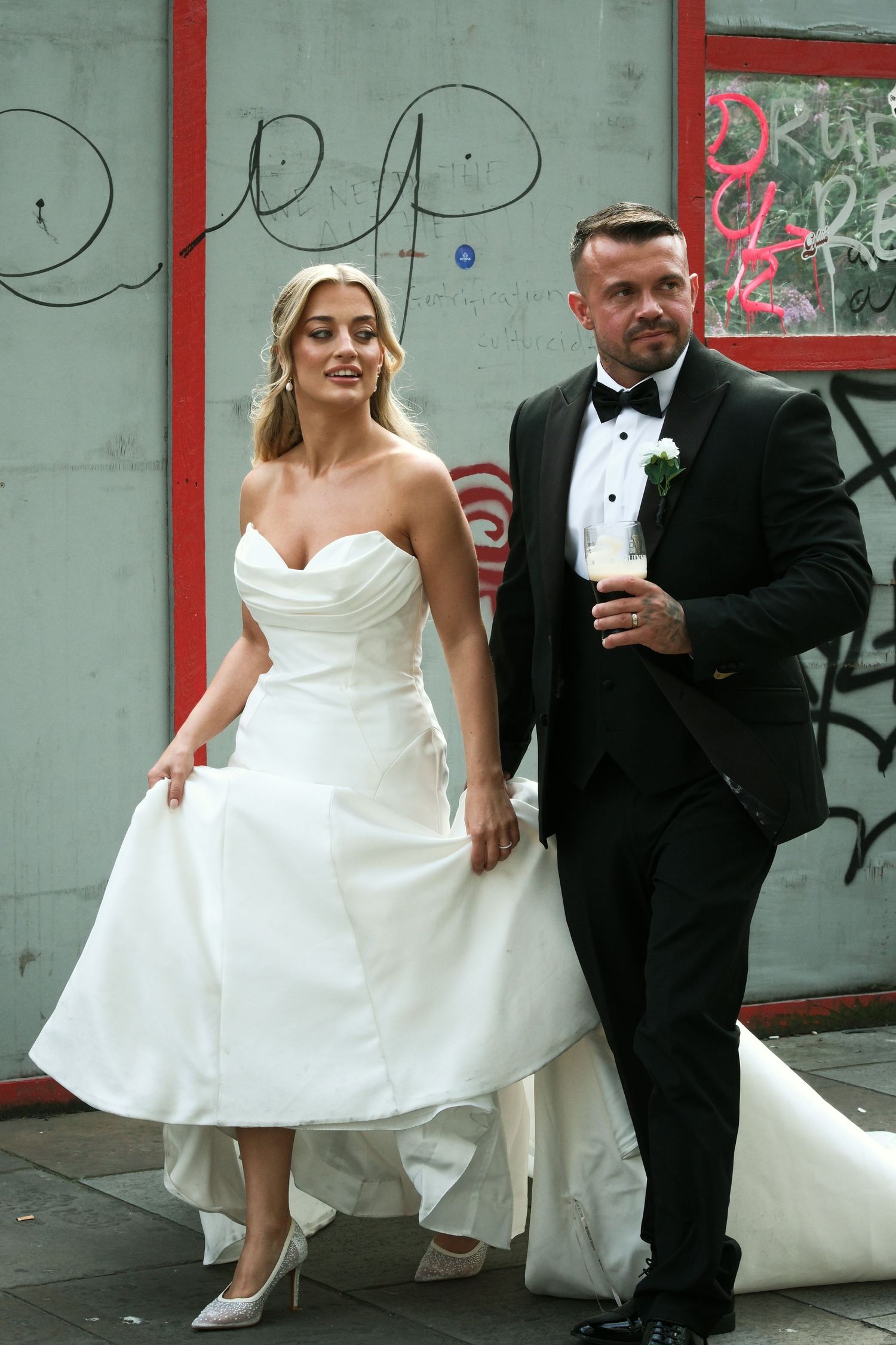 A bride in a white strapless gown and groom in black tuxedo pose together in front of a graffiti wall.