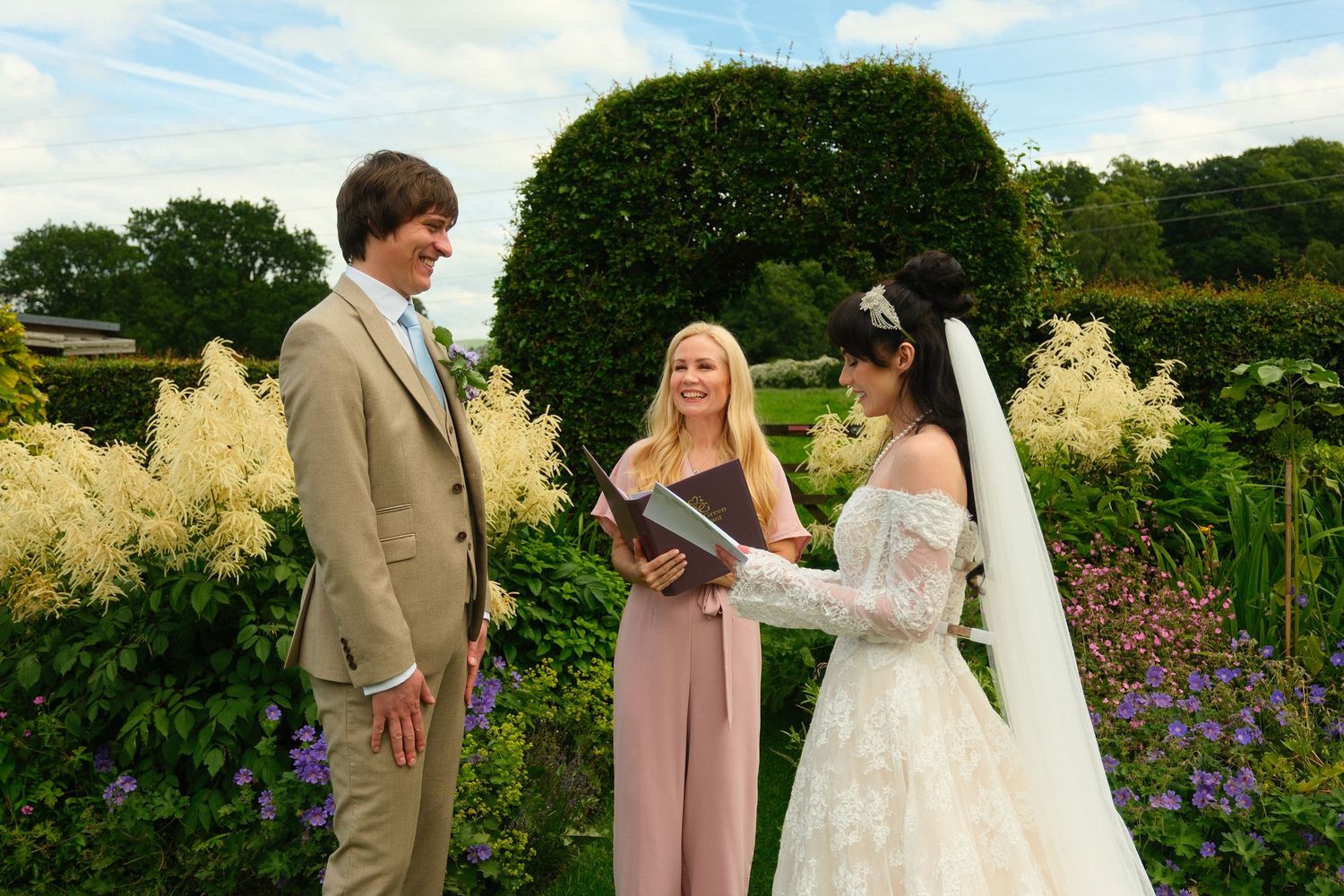 Bride and groom exchange vows with female officiant in lush garden ceremony setting.