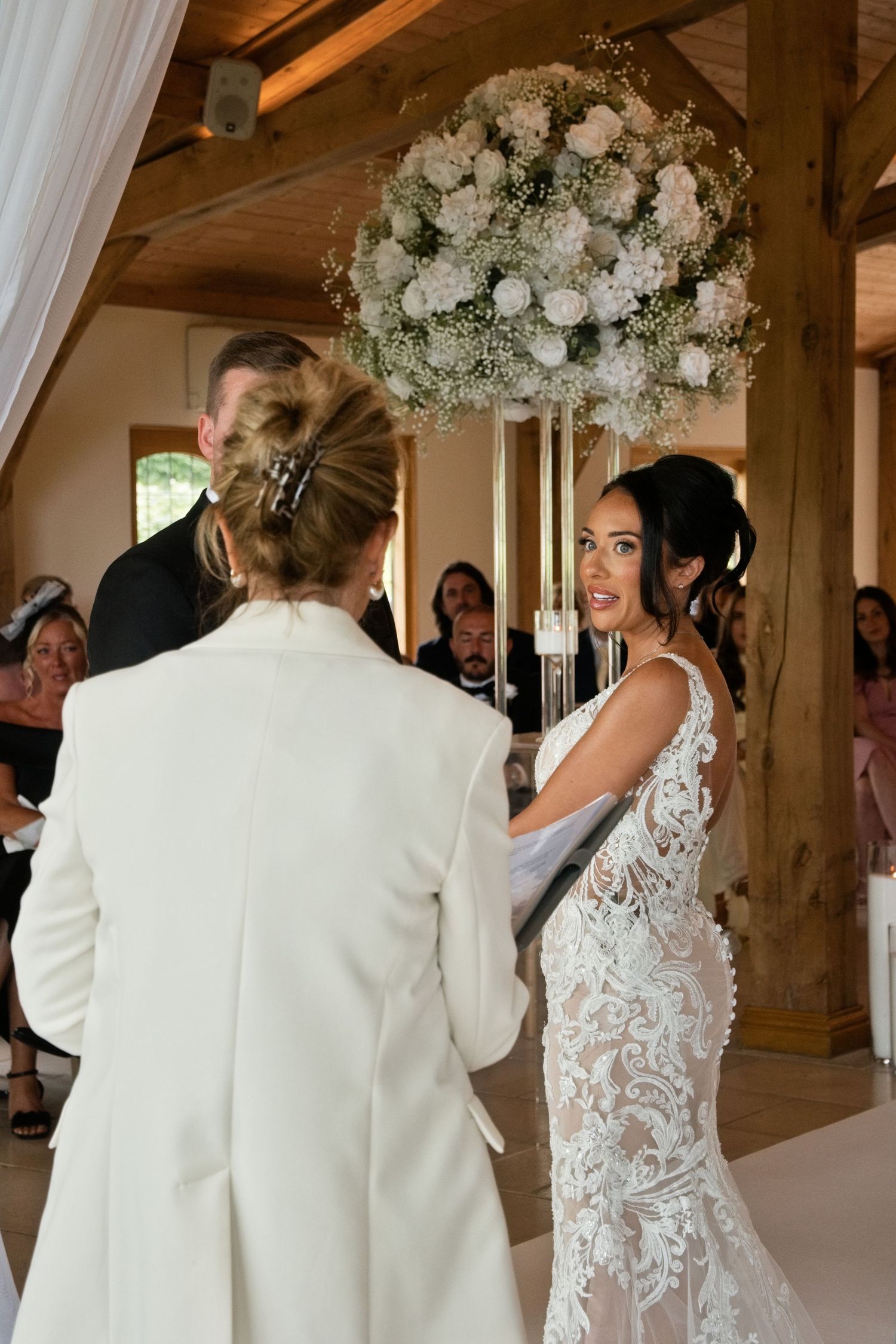 Bride in intricate lace gown exchanges vows with officiant in white suit under large white floral arrangement.
