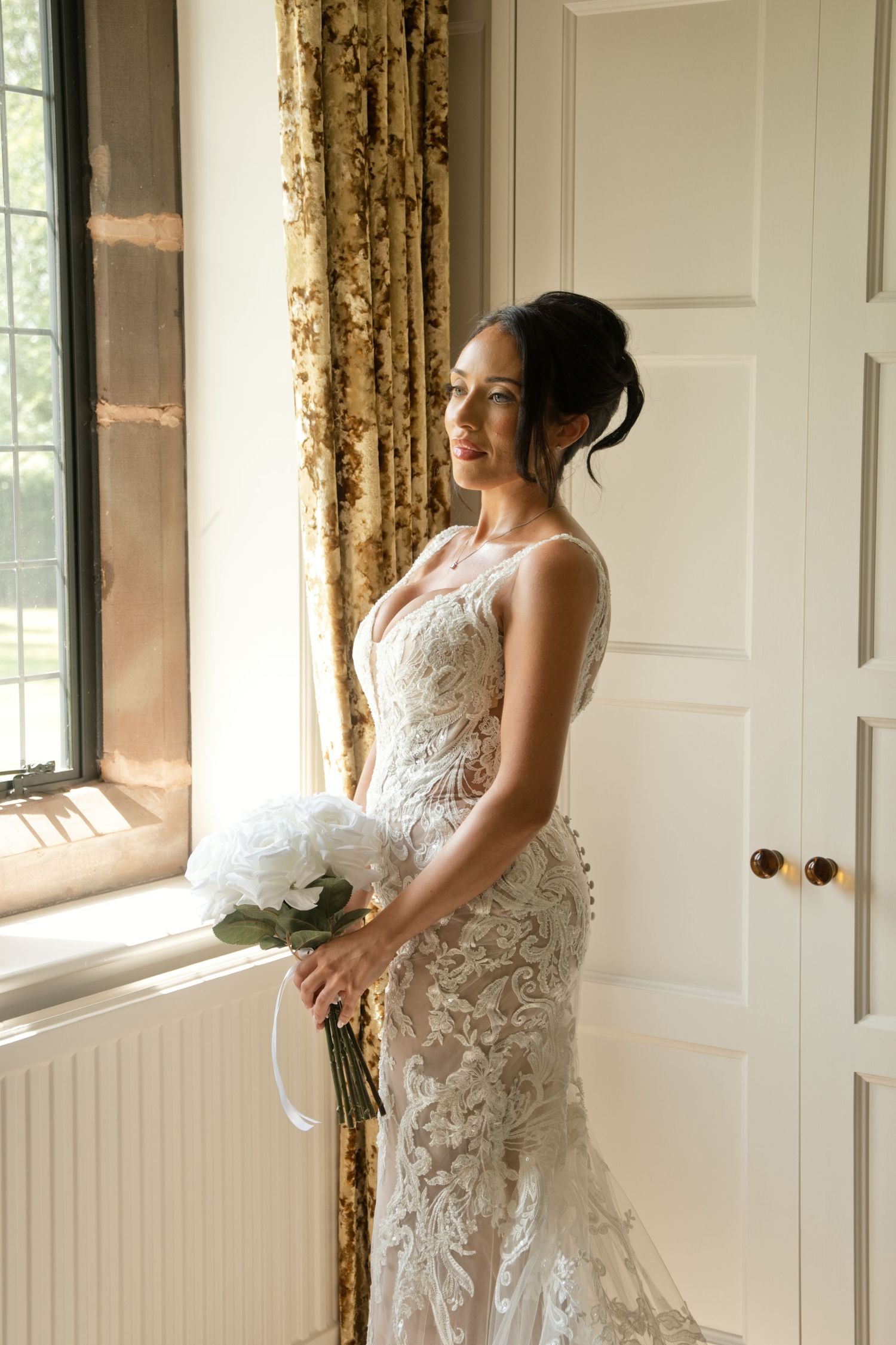 Elegant bride in lace mermaid wedding gown holds white bouquet while gazing out sunlit window with gold curtains.