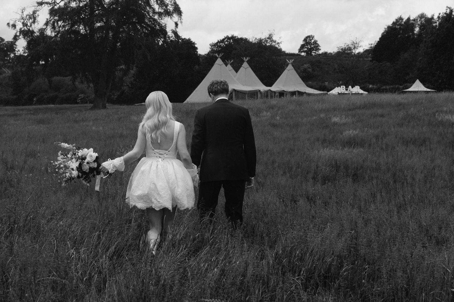 Bride and groom walk hand-in-hand through green meadow toward elegant tipi wedding tents in black and white photo.