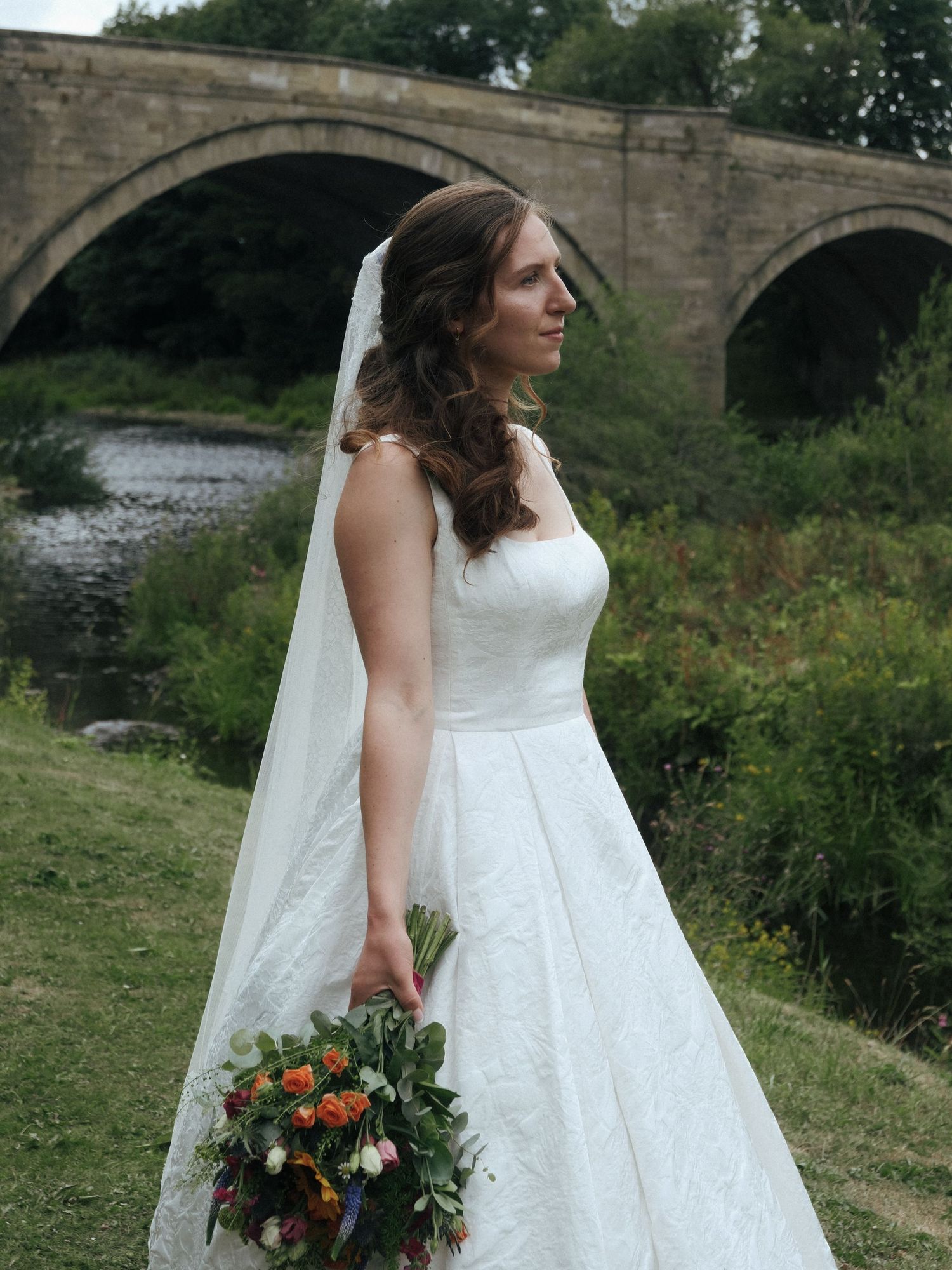 Bride in white lace gown holding colorful bouquet stands before a stone arch bridge over a river.