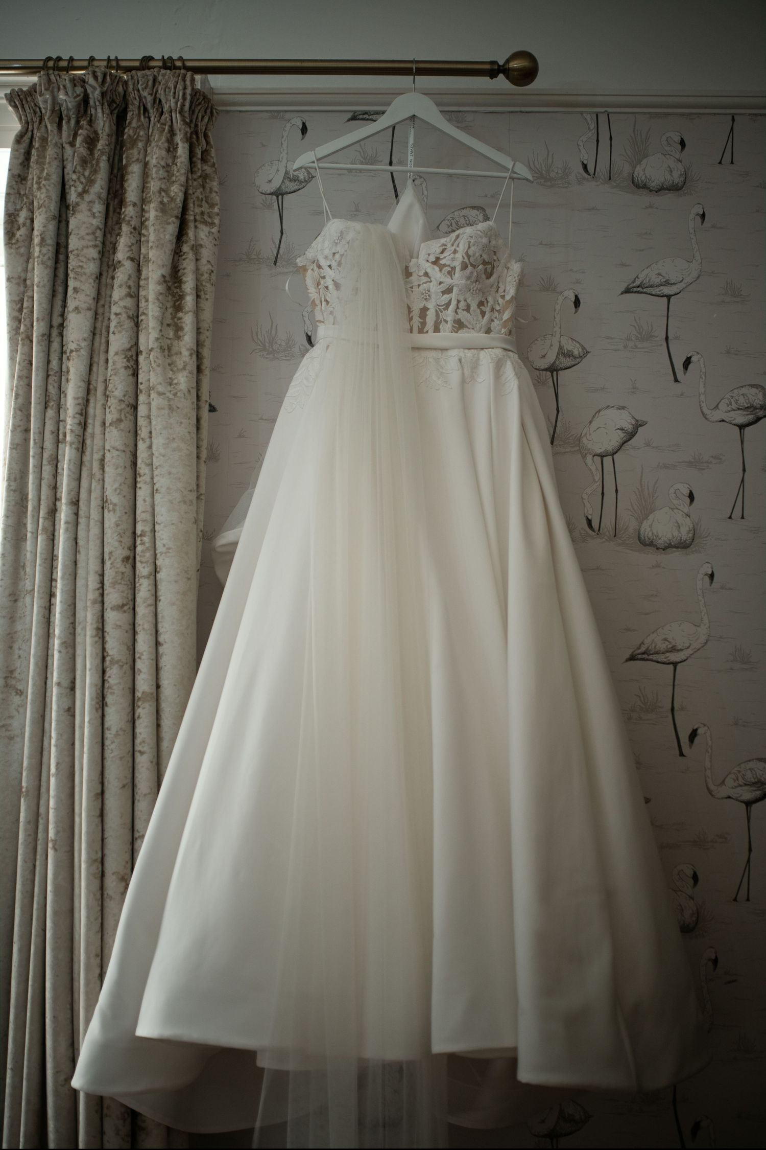 Elegant white lace A-line wedding dress hanging on a hanger against floral wallpaper.