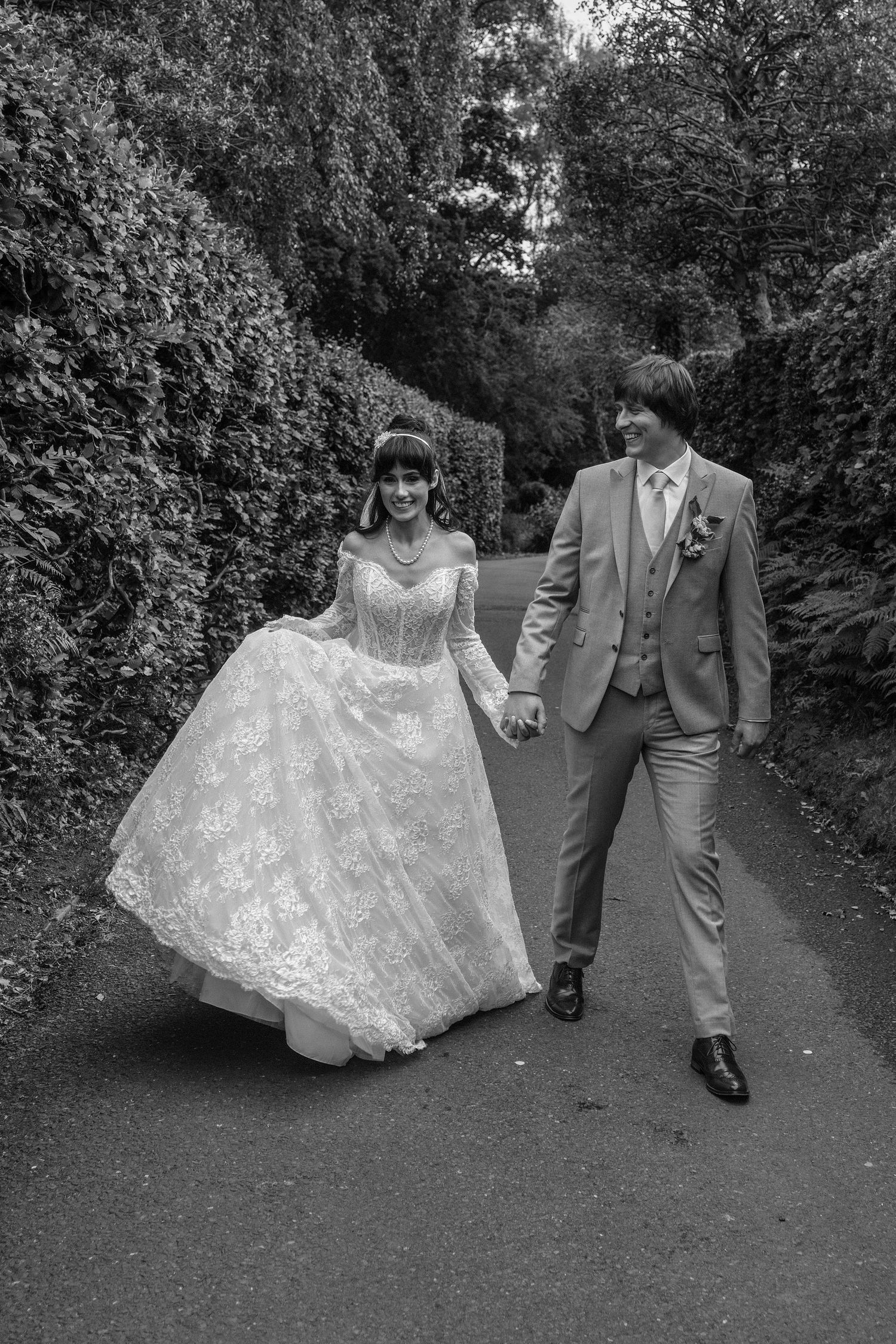 Black and white photo of smiling bride and groom walking hand in hand along tree-lined garden path.