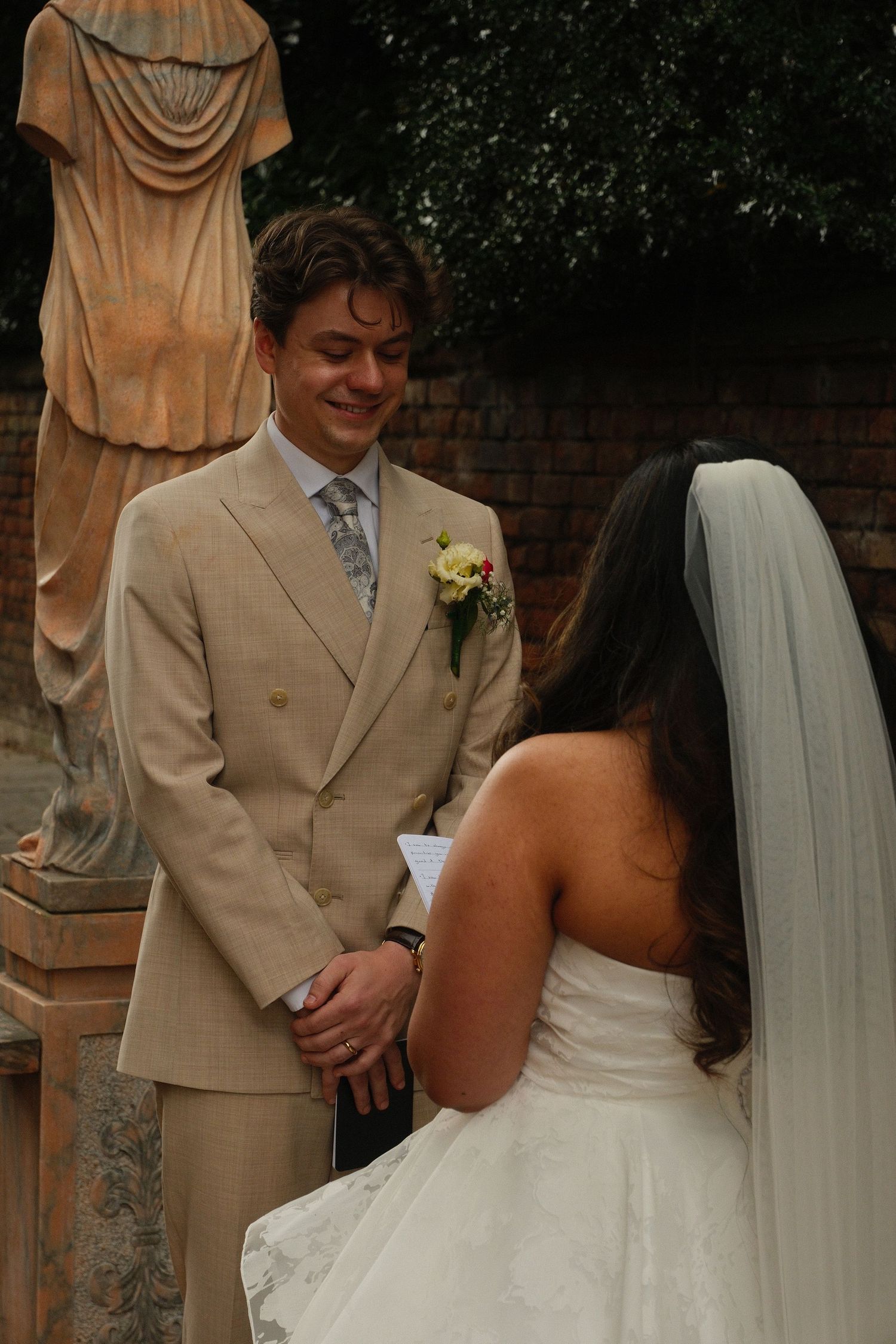 Smiling groom in beige suit faces bride in white strapless gown during outdoor wedding ceremony with stone statues.