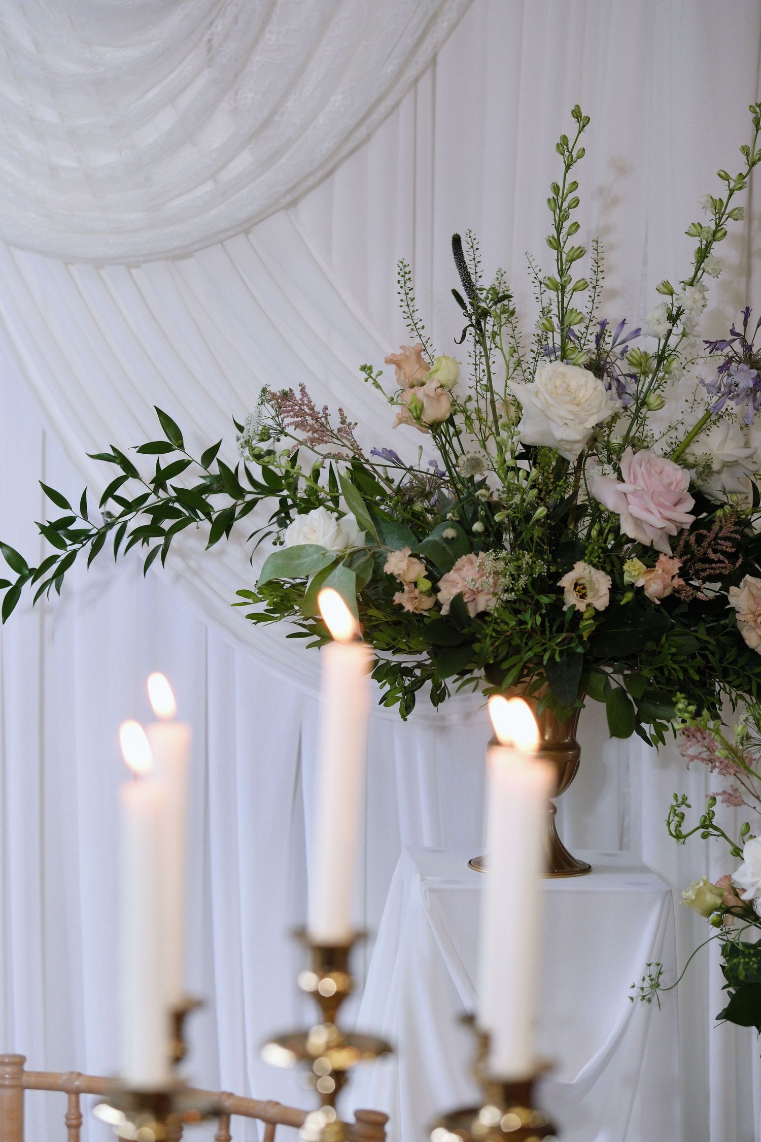 Elegant wedding decor with lit white candles on gold candelabra and a floral arrangement of roses and greenery against white draping.