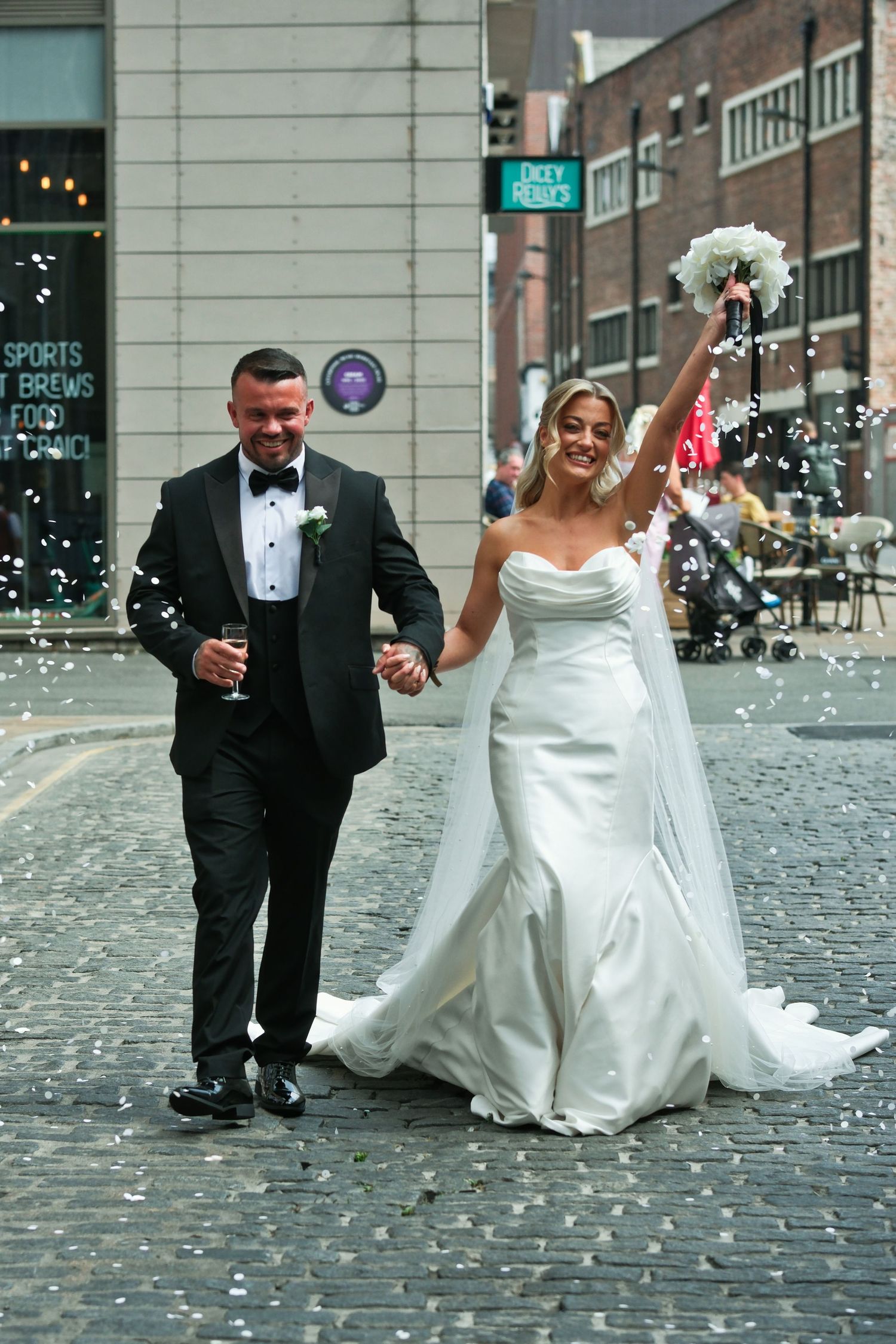 Happy bride and groom walking through confetti shower on cobblestone street after wedding.