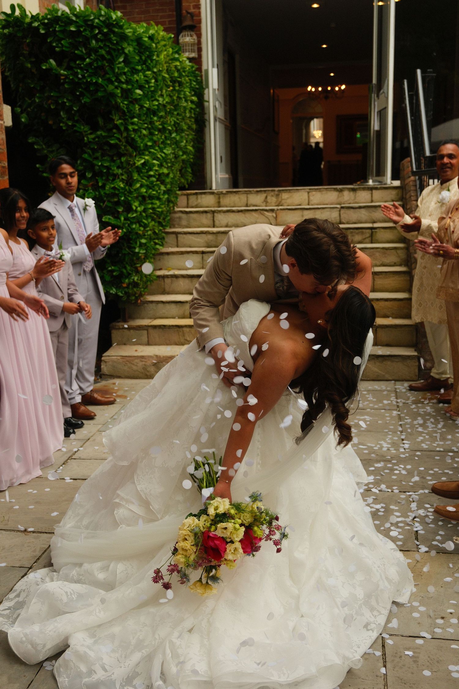 Groom dips bride for a romantic kiss as confetti falls, surrounded by applauding guests on wedding day.
