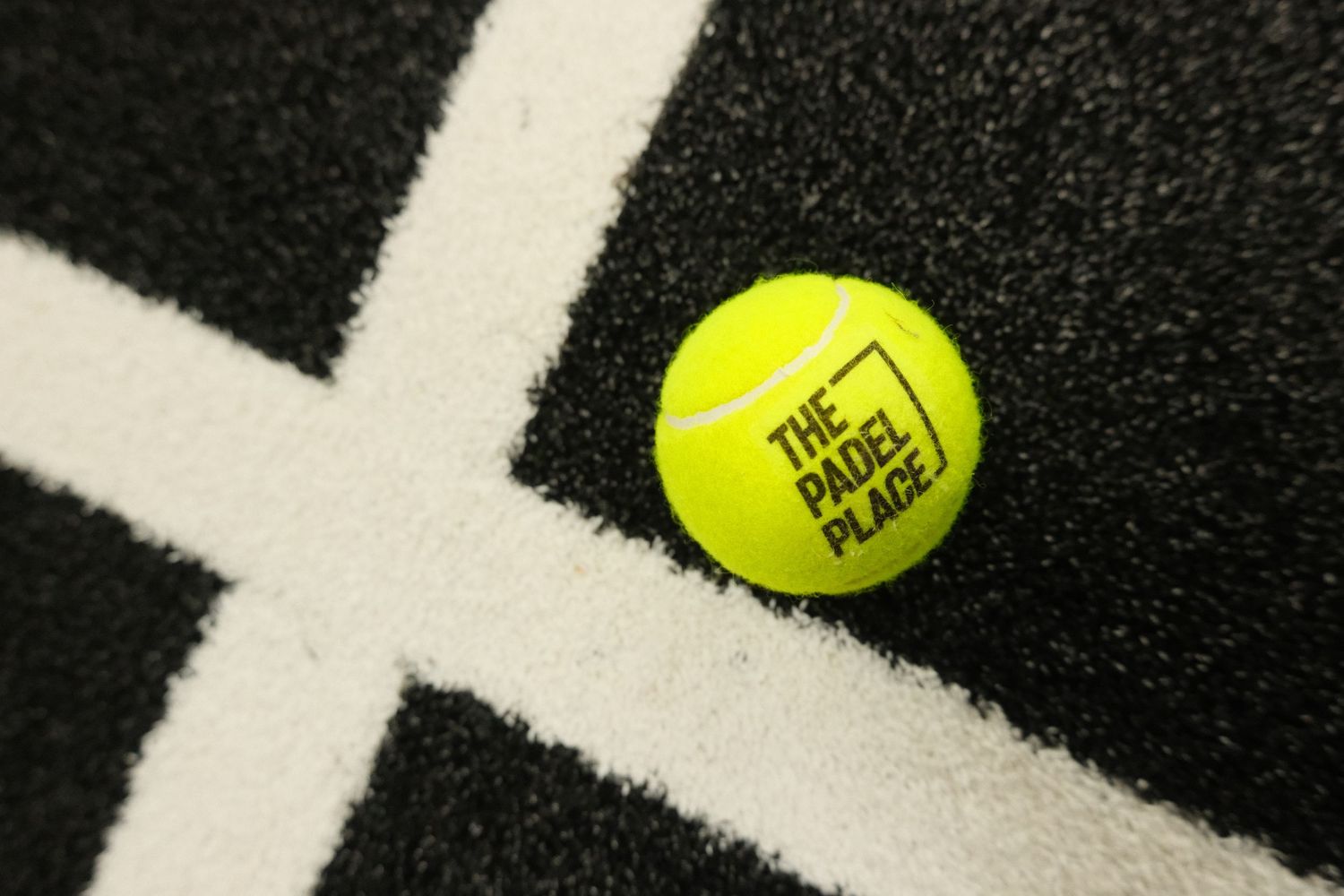 A yellow tennis ball with The Padel Place logo resting on a dark padel court surface with white lines.