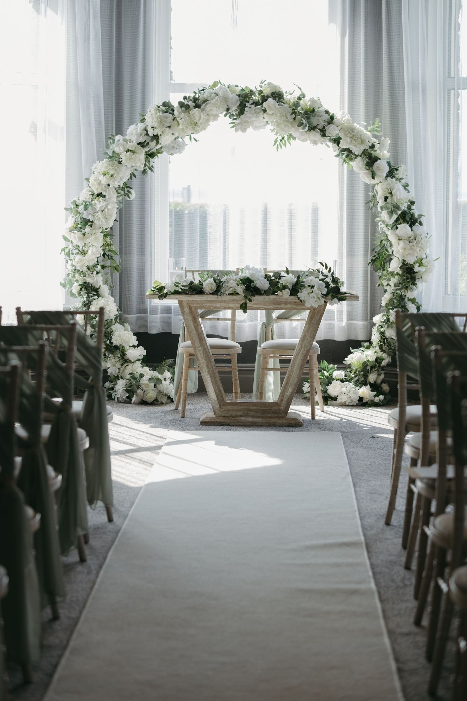 Elegant wedding ceremony setup with white floral arch, wooden chairs, and a white aisle runner in a bright indoor venue.