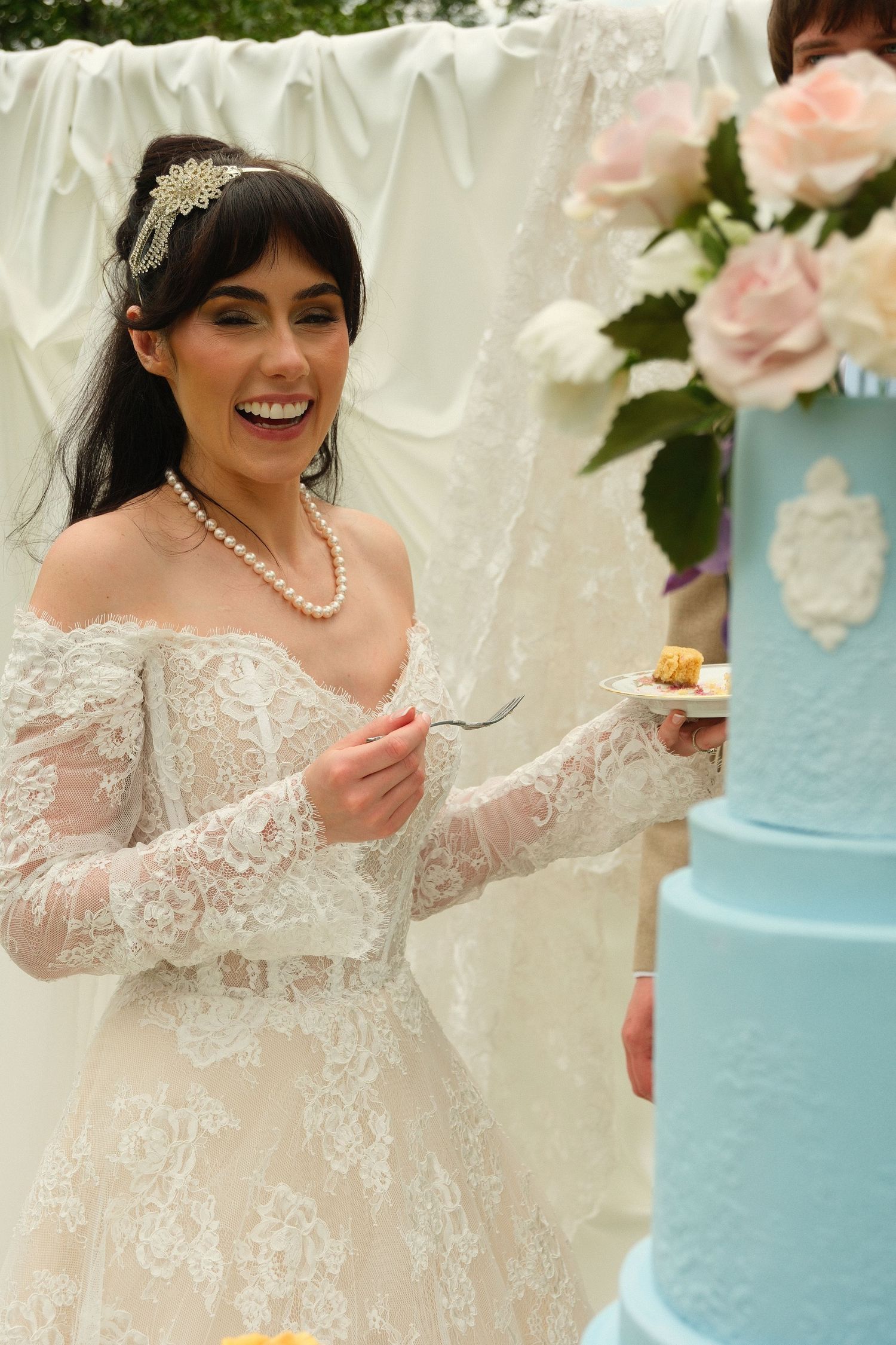 Smiling bride in lace gown and pearls enjoys wedding cake slice beside blue tiered cake.