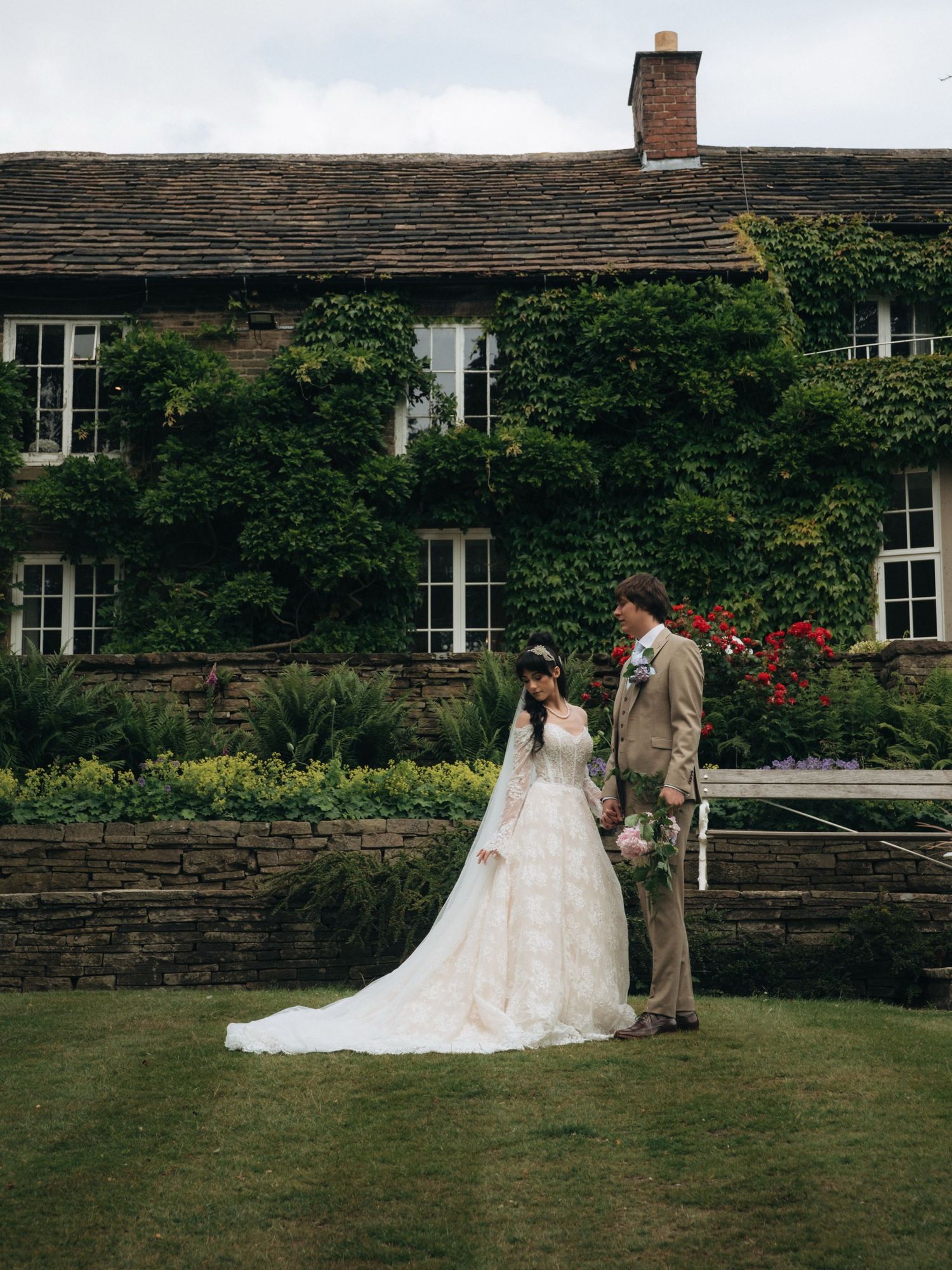 Bride and groom stand on lush lawn before ivy-covered stone manor, bride in flowing white gown with long veil.