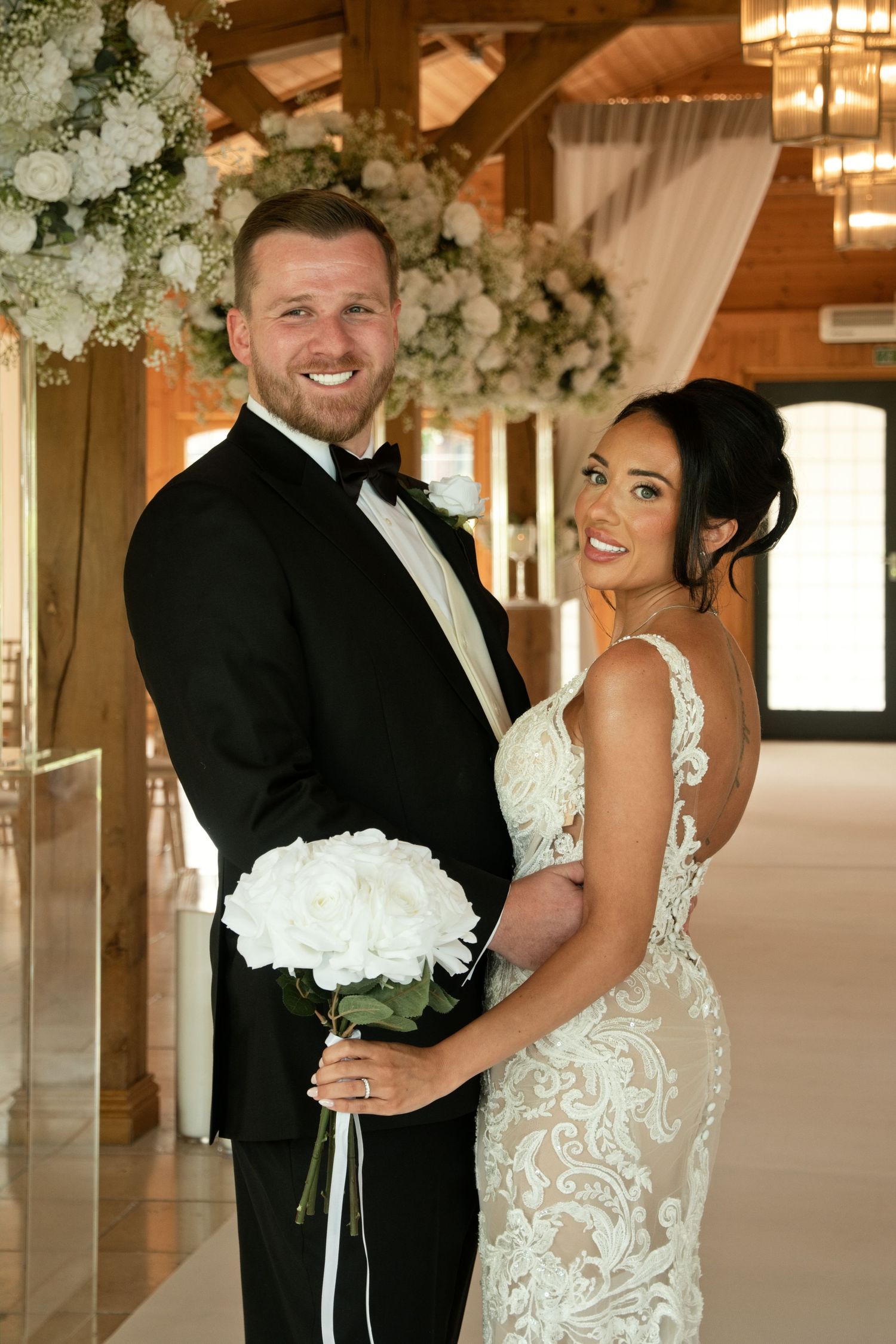 Smiling bride and groom pose together indoors, she holds white peony bouquet, wearing lace gown and black tuxedo.