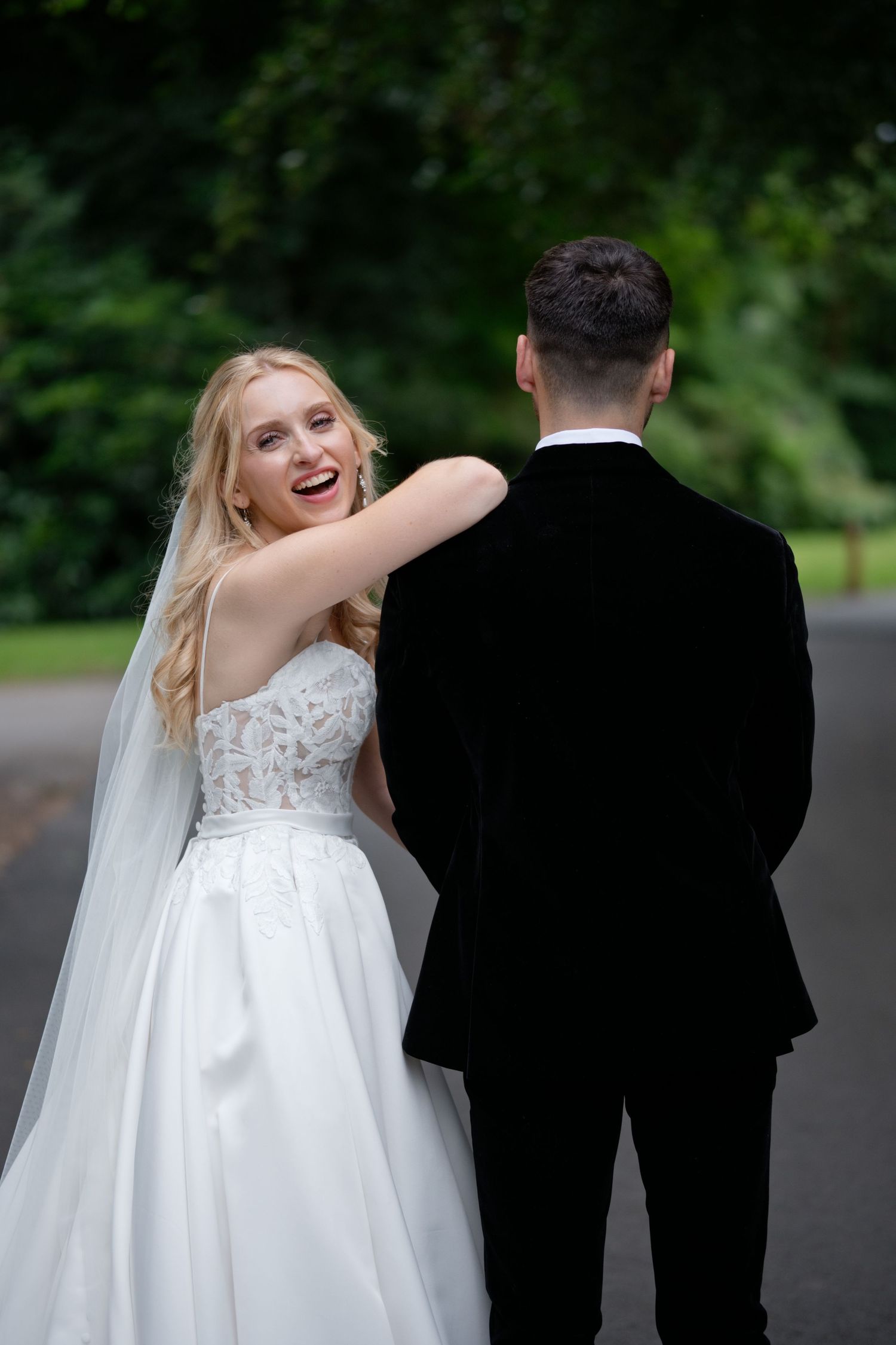 Laughing bride in white lace gown looks back at groom while walking together on a tree-lined path.