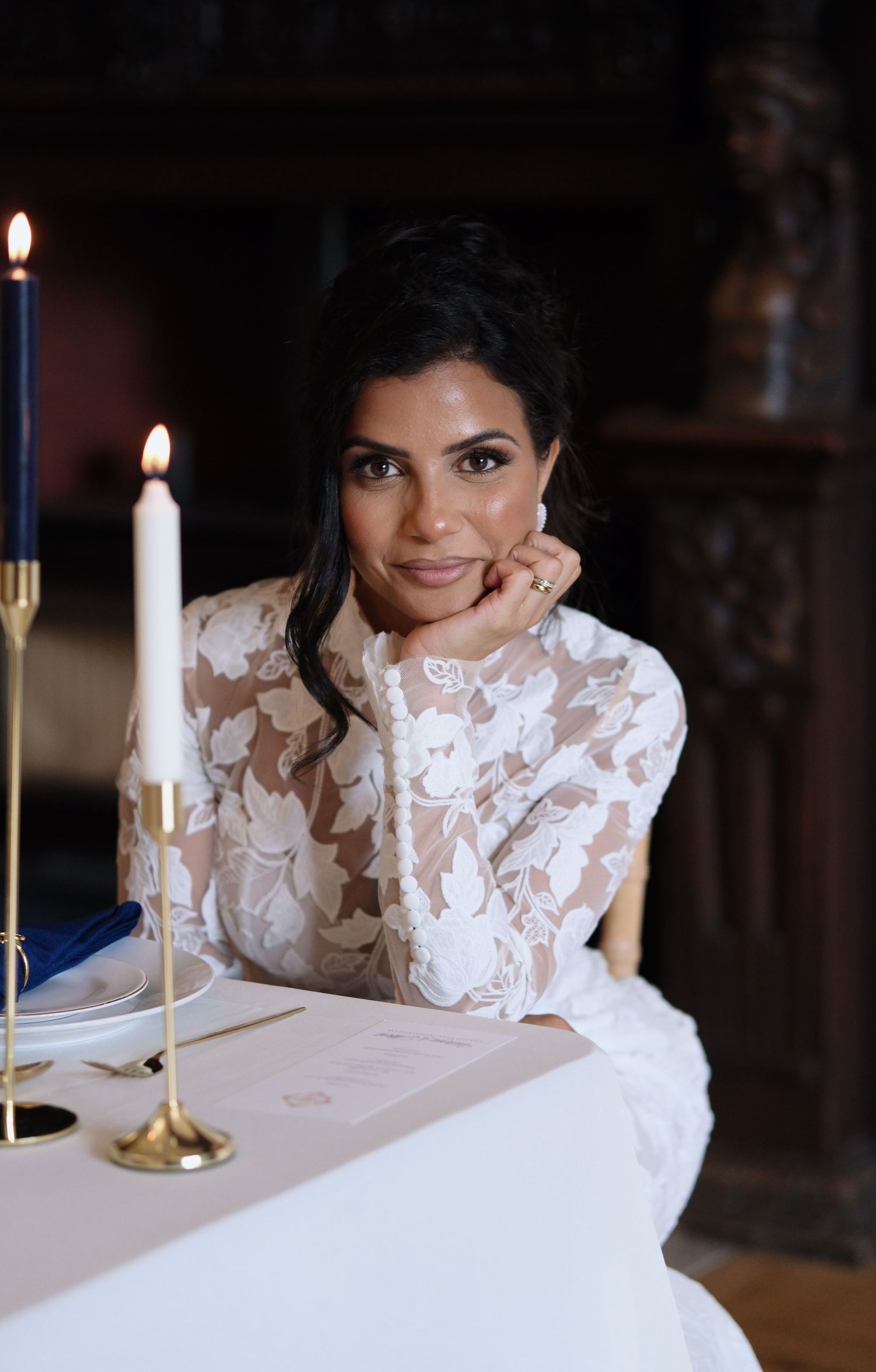 Elegant bride in white lace gown seated at candlelit table with gold candlesticks and blue napkins.