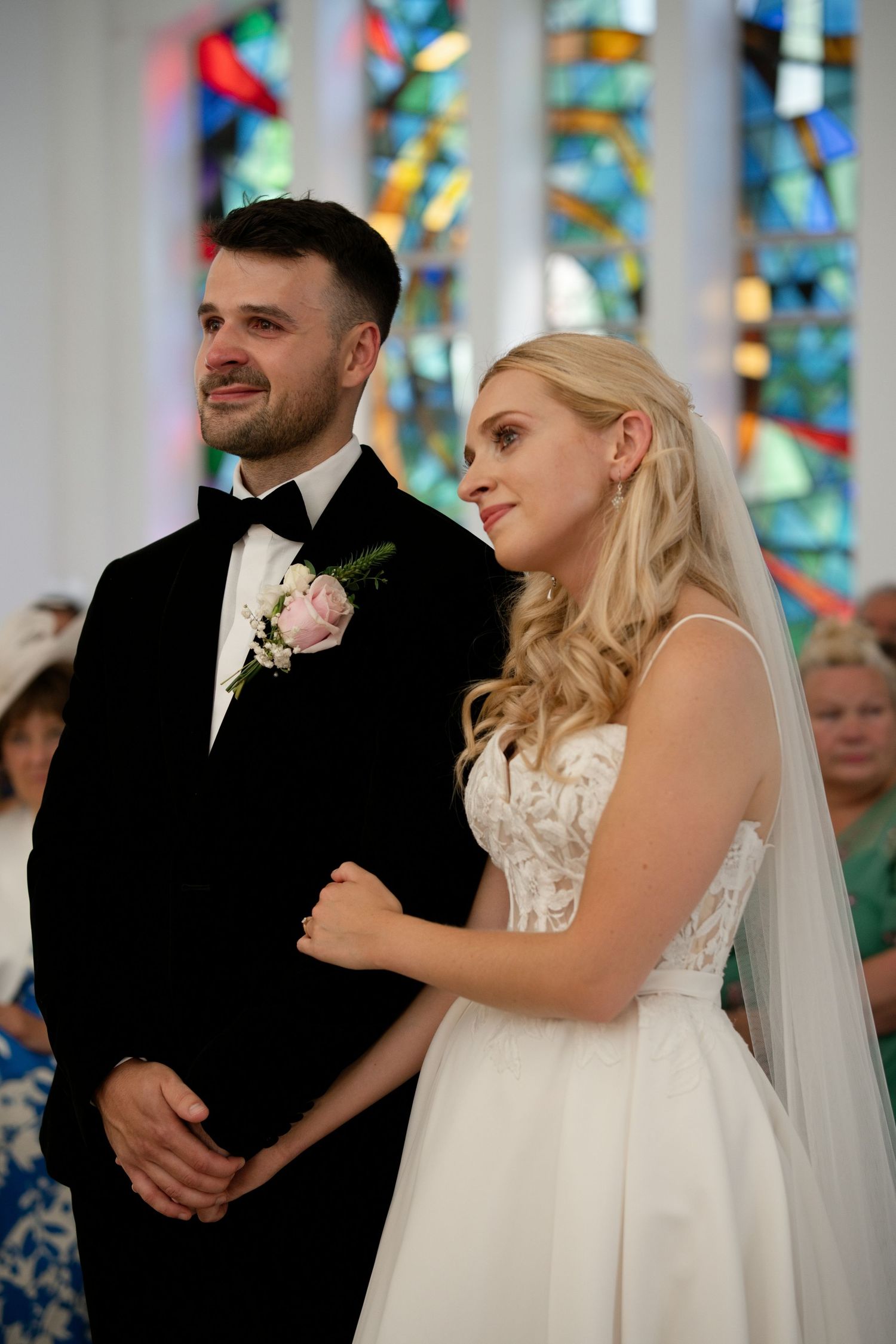 Smiling bride in white gown and groom in tuxedo stand together during church wedding ceremony.