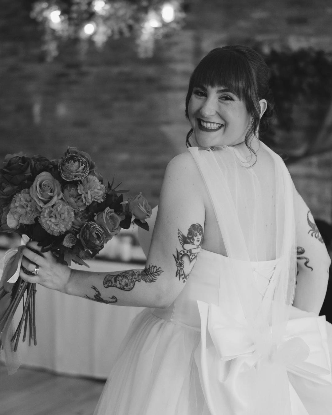 Black and white photo of tattooed bride laughing while holding colorful floral bouquet in white gown.