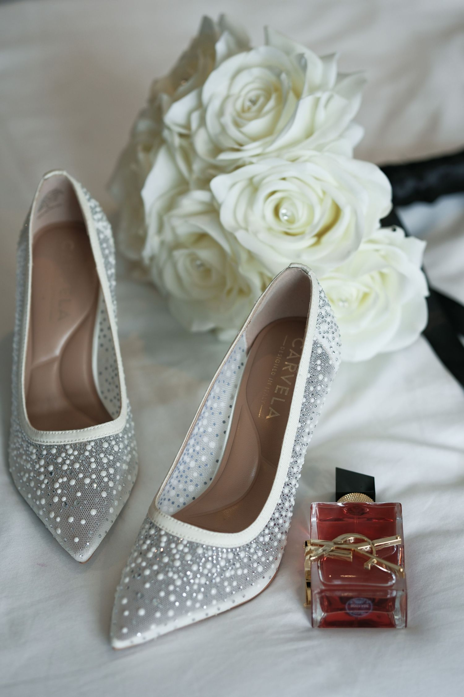 Sparkling crystal-embellished bridal heels beside white roses bouquet and red YSL perfume bottle on white fabric.