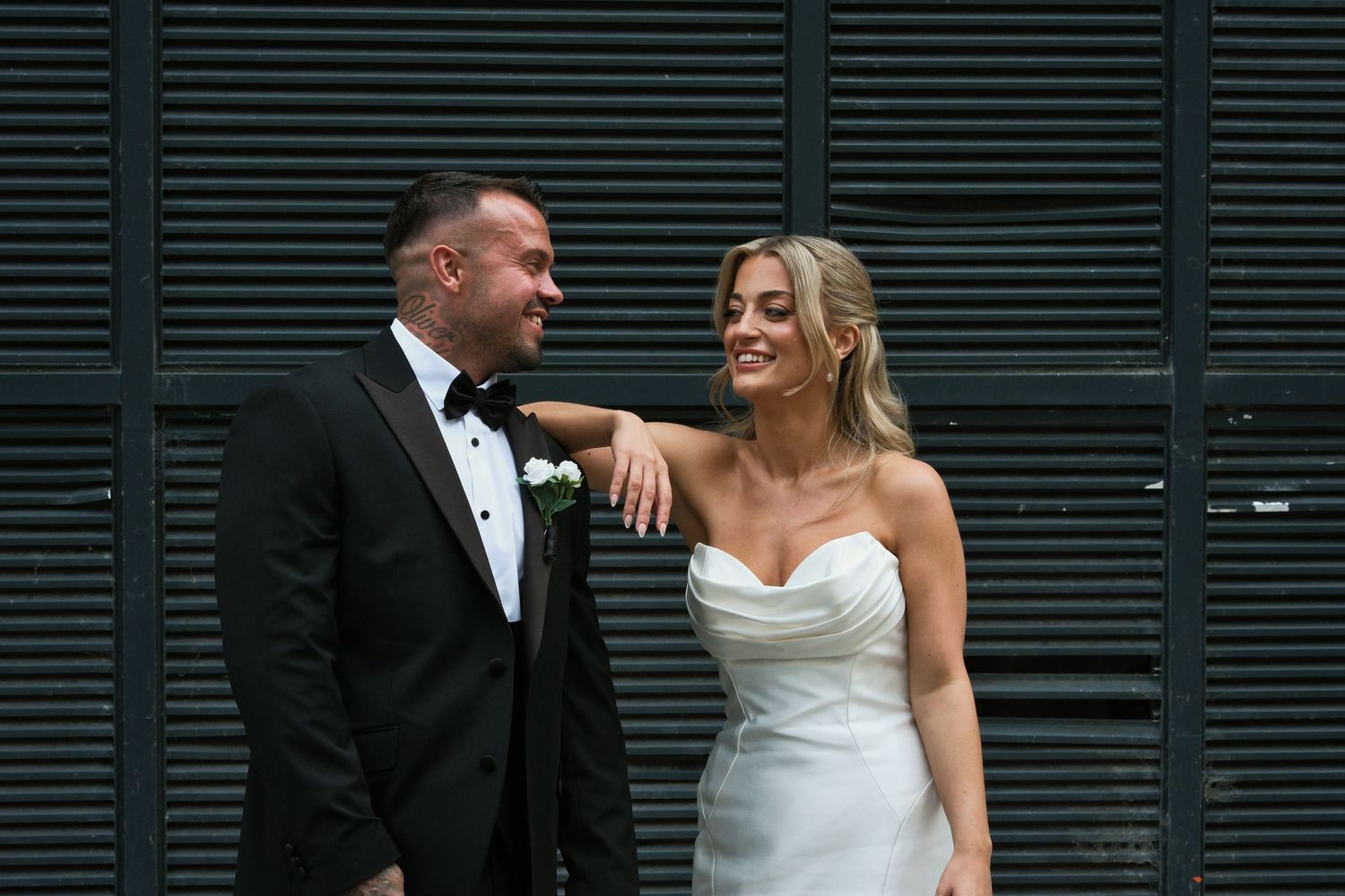 Bride in strapless white gown and groom in black tuxedo smile at each other outdoors.