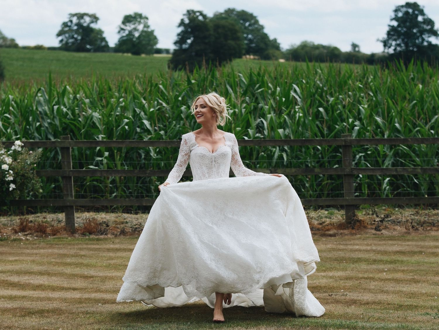 Bride in a long-sleeve white lace wedding gown twirling on green grass with a cornfield and wooden fence backdrop.