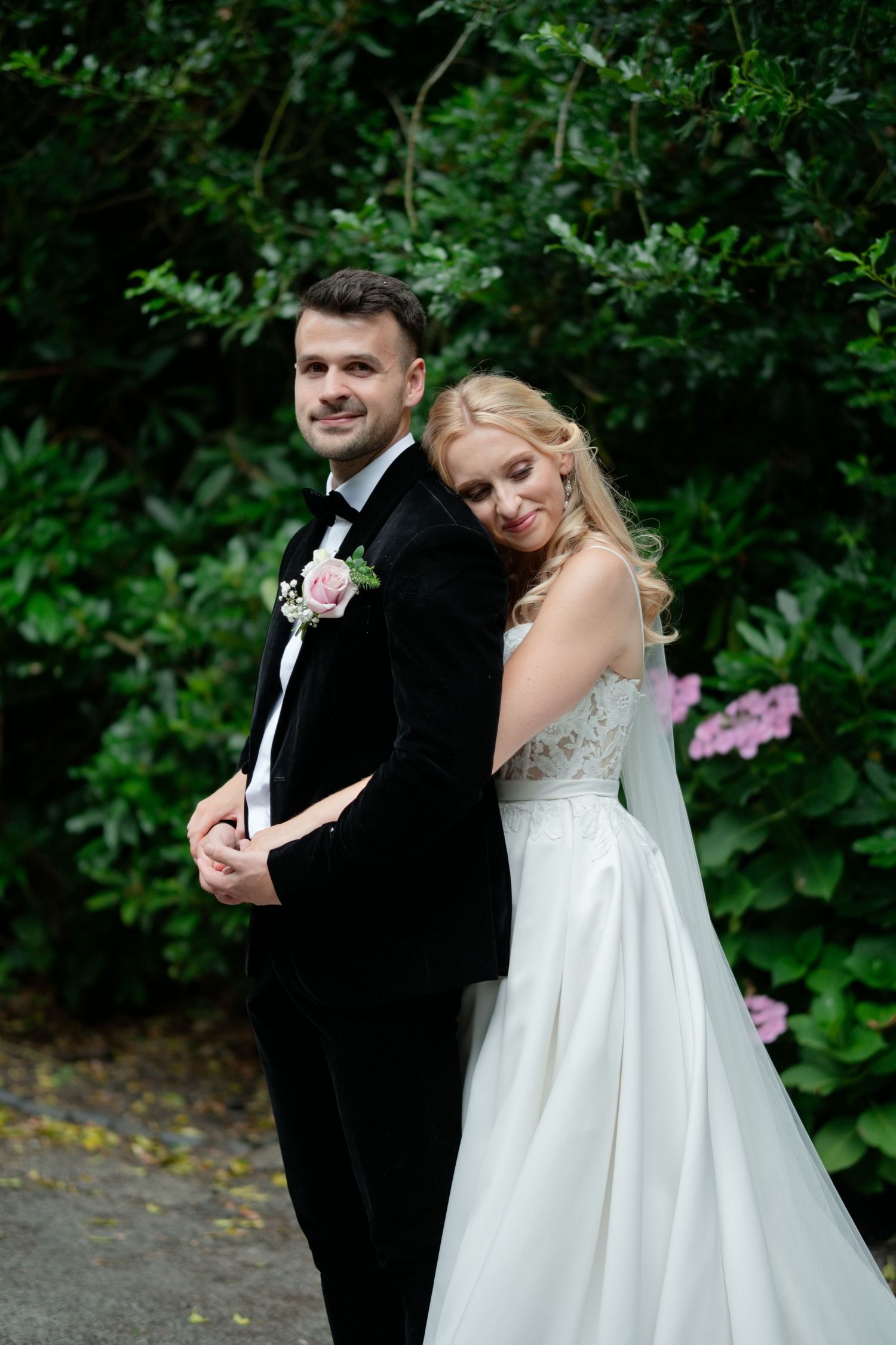 Smiling groom in black tuxedo with bride in white lace wedding dress embracing outdoors amid green foliage.