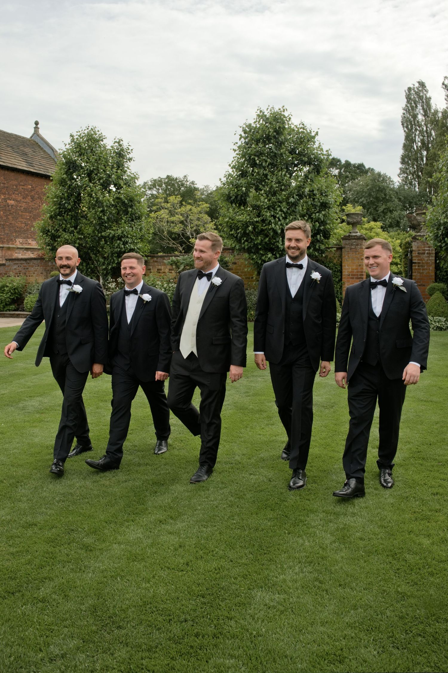 Five groomsmen in black tuxedos walk together laughing across a manicured lawn at an outdoor wedding venue.