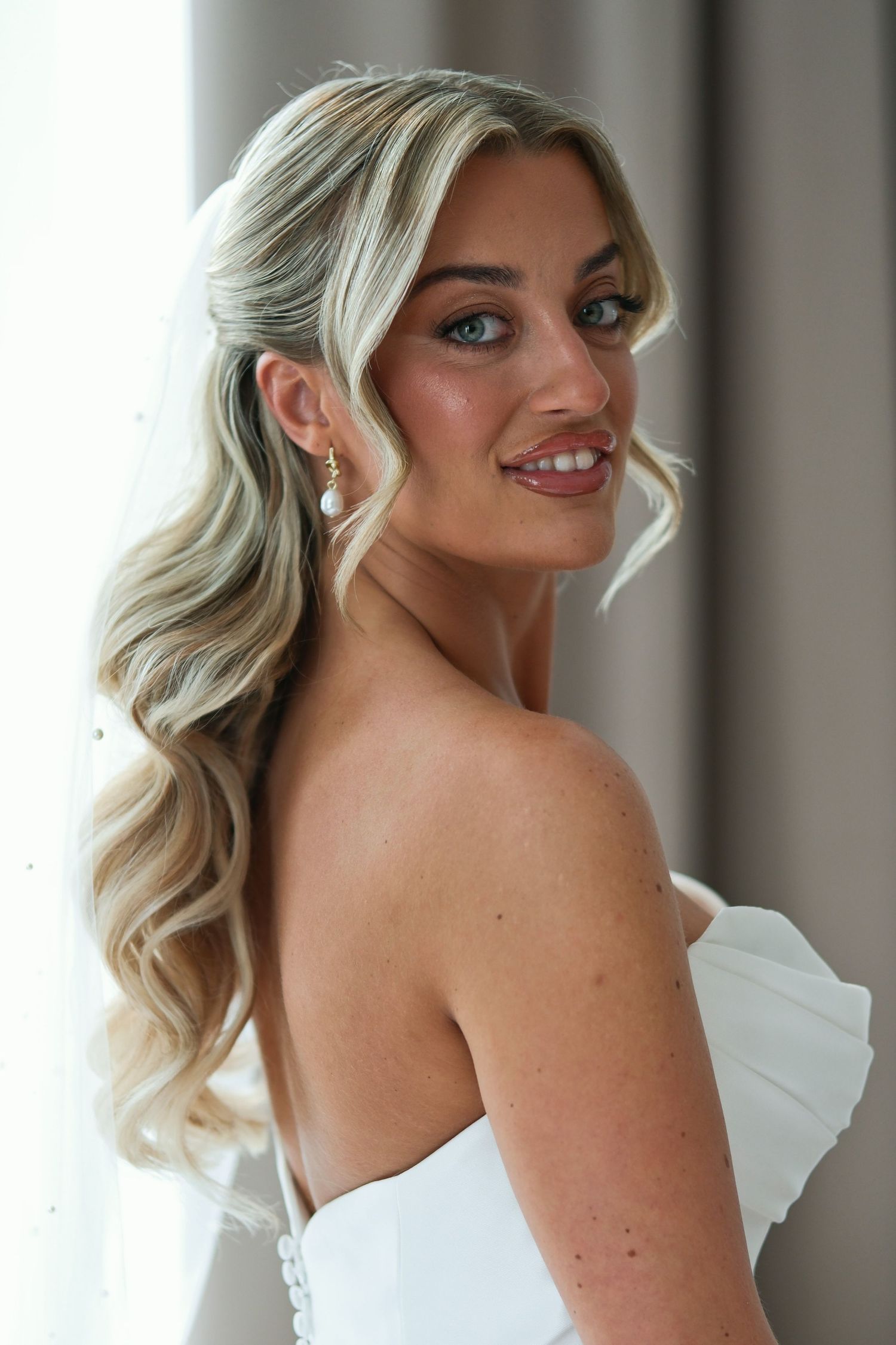 Smiling bride with blonde wavy hair, pearl earrings, and a white strapless gown, posing elegantly near a window.