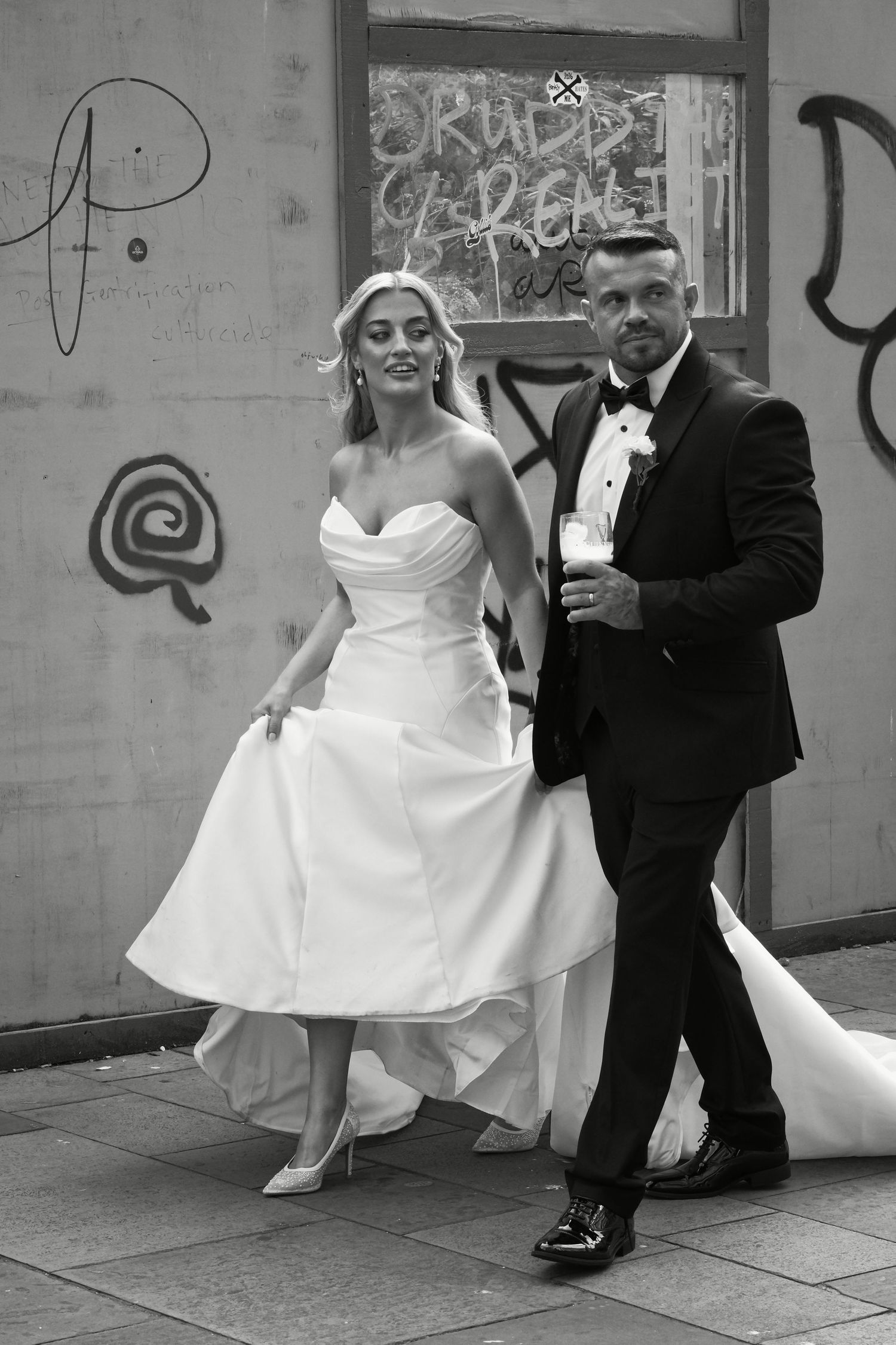 Black and white photo of bride and groom walking past graffiti wall on their wedding day.