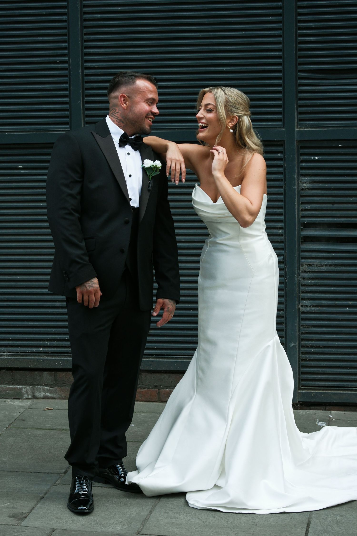 Laughing bride and groom posing together against dark metal wall, bride in fitted white gown.