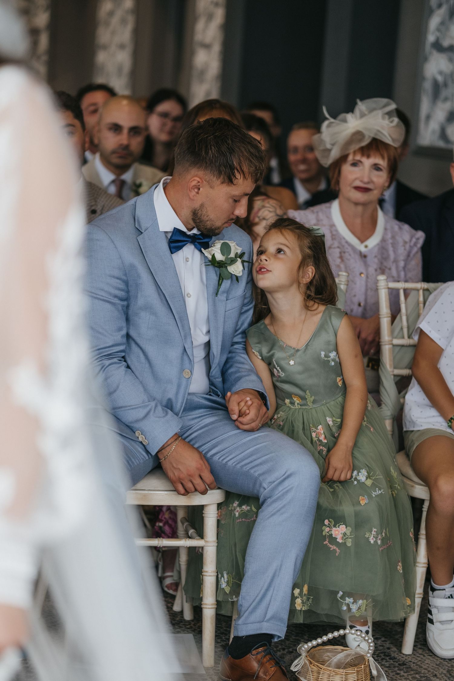 Groom in light blue suit shares tender moment with flower girl in green floral dress during outdoor wedding ceremony.