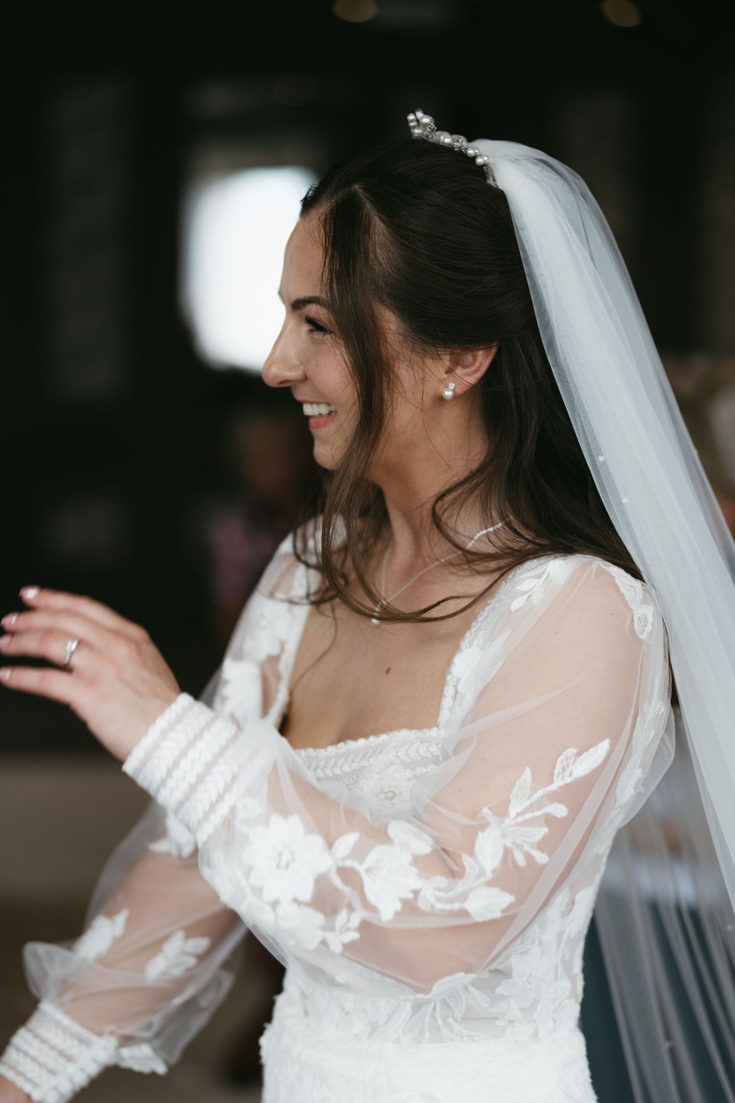 Smiling bride in lace long-sleeve wedding dress and veil, wearing pearl earrings and tiara.